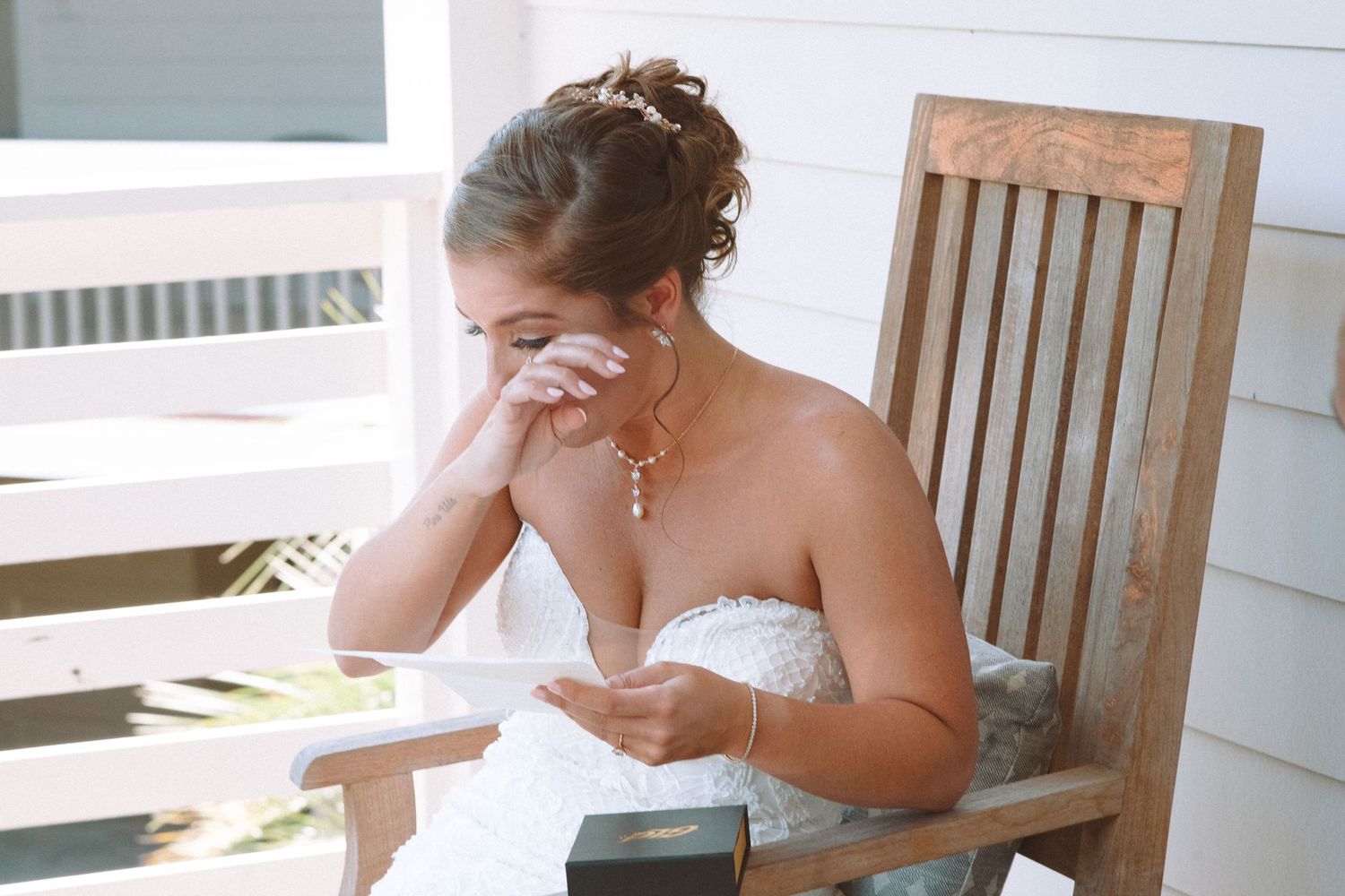 Emotional bride in strapless lace gown wiping tears while reading a letter on wooden chair.