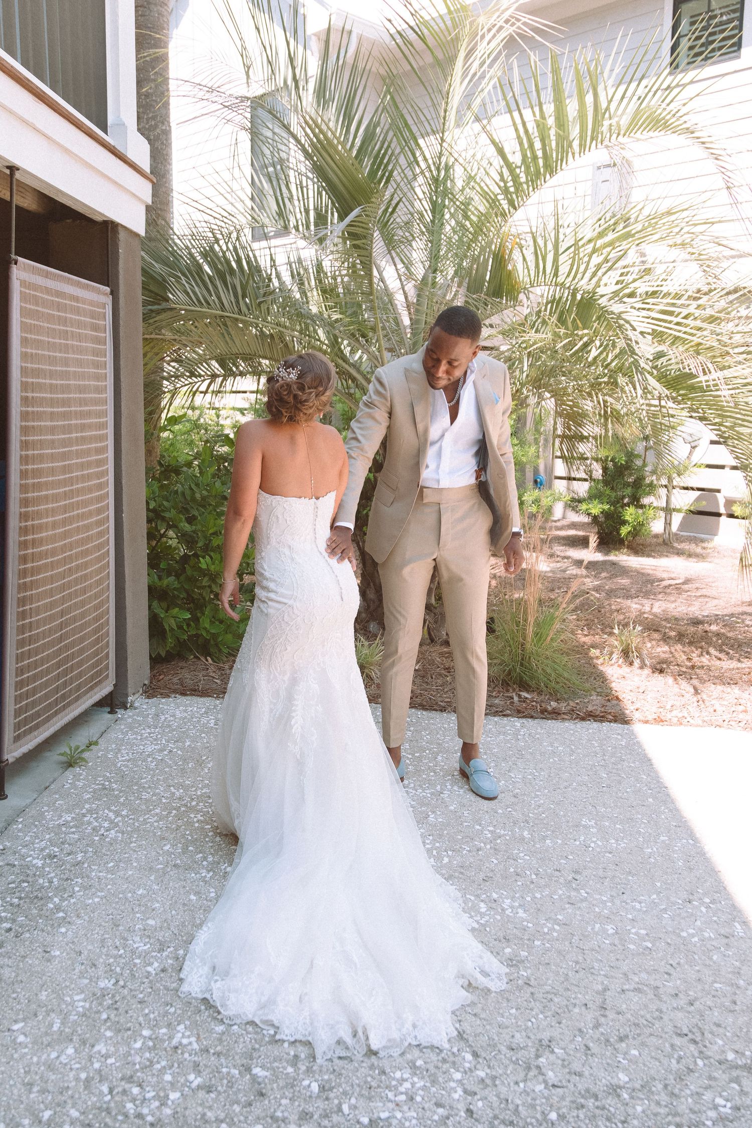 Bride in lace mermaid gown and groom in tan suit walk together hand in hand near tropical palms.