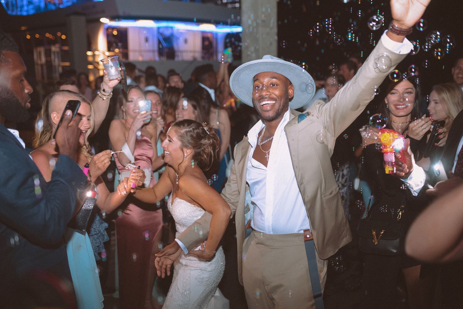 Groom in tan suit and wide-brim hat raises hand while dancing with bride at festive wedding reception.