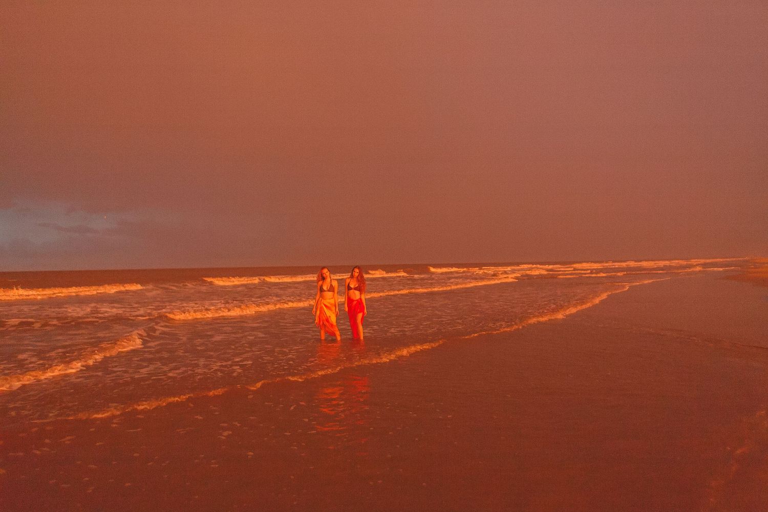 Two people walking along a beach at sunset with vivid orange sky.