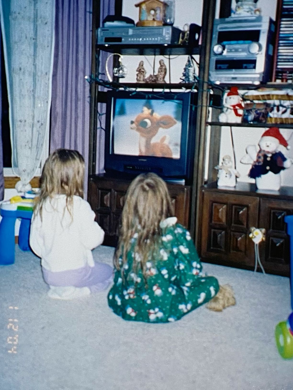 Two young girls sitting on the floor watching a deer cartoon on a living room TV.