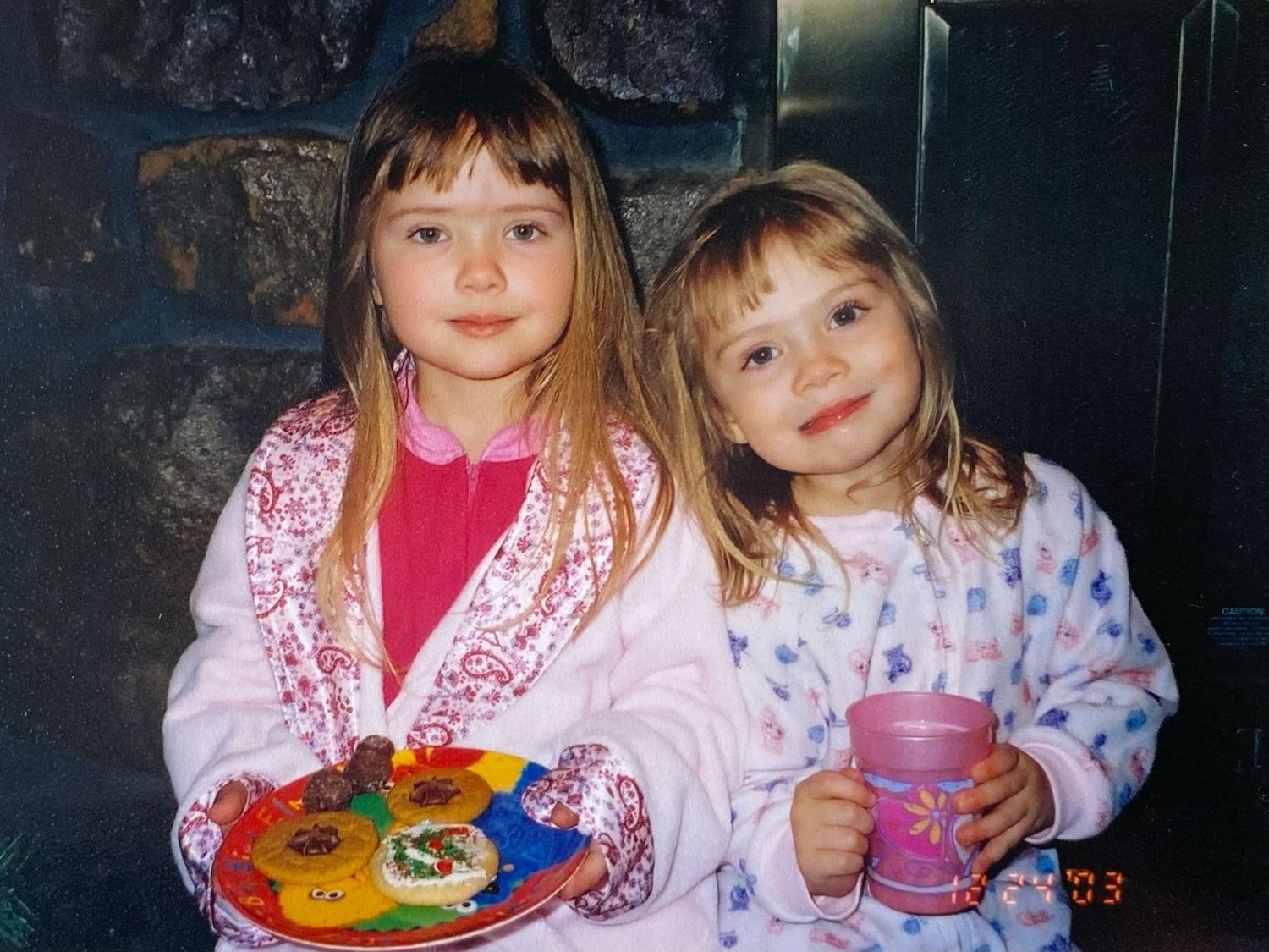 Two young girls in pajamas smiling while holding a plate of decorated holiday cookies and a pink cup.