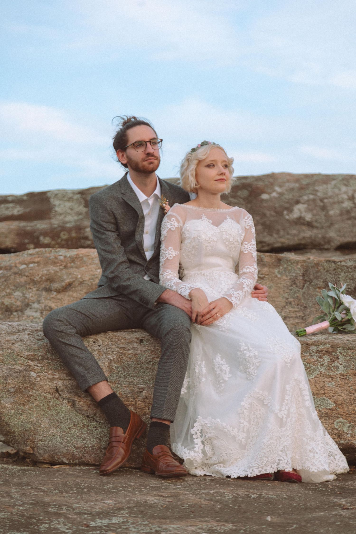 A bride in lace gown and groom in gray suit sit together on rocks outdoors on their wedding day.