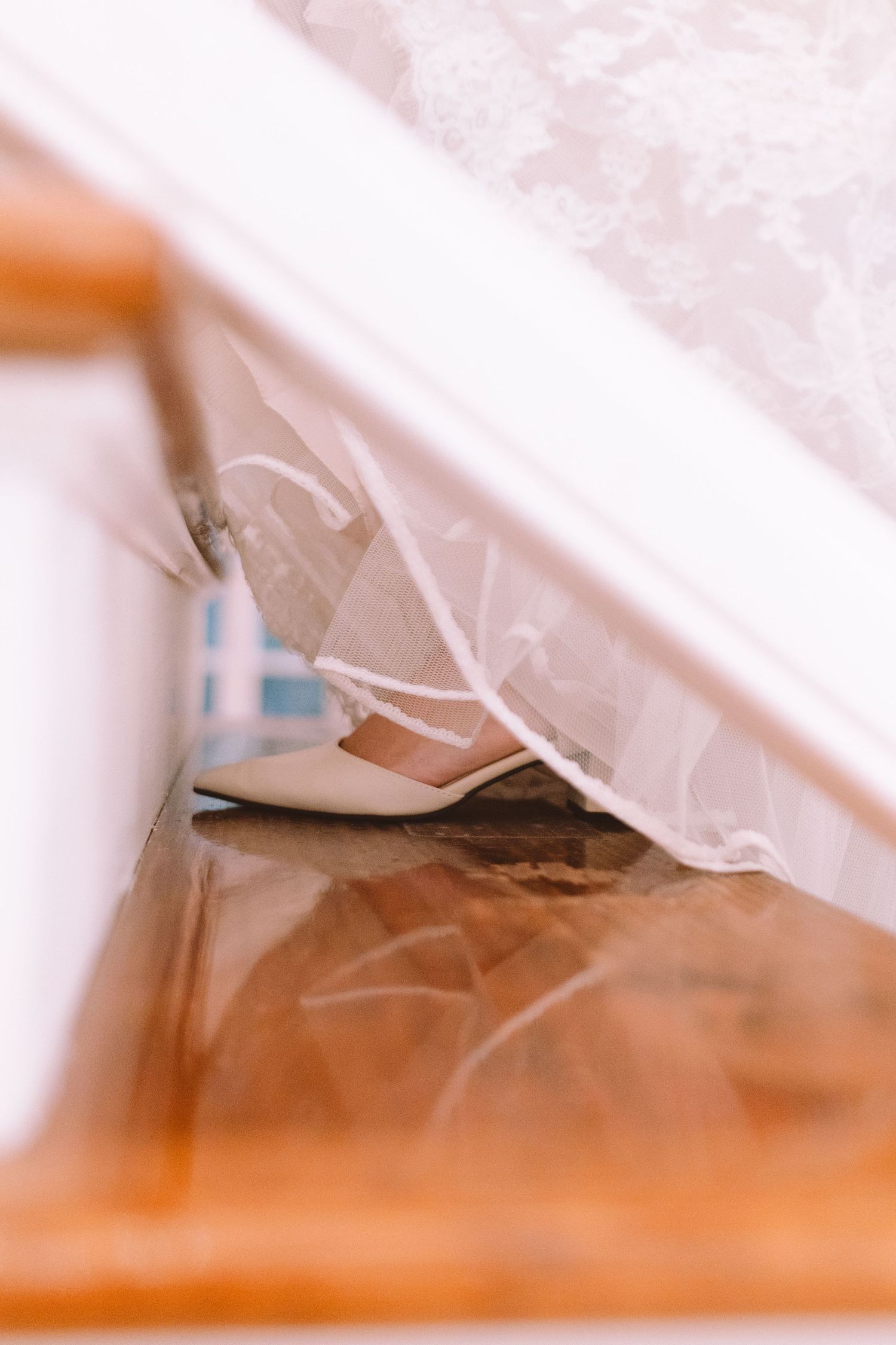 Close-up of brides ivory wedding shoe peeking beneath lace gown on wooden floor.