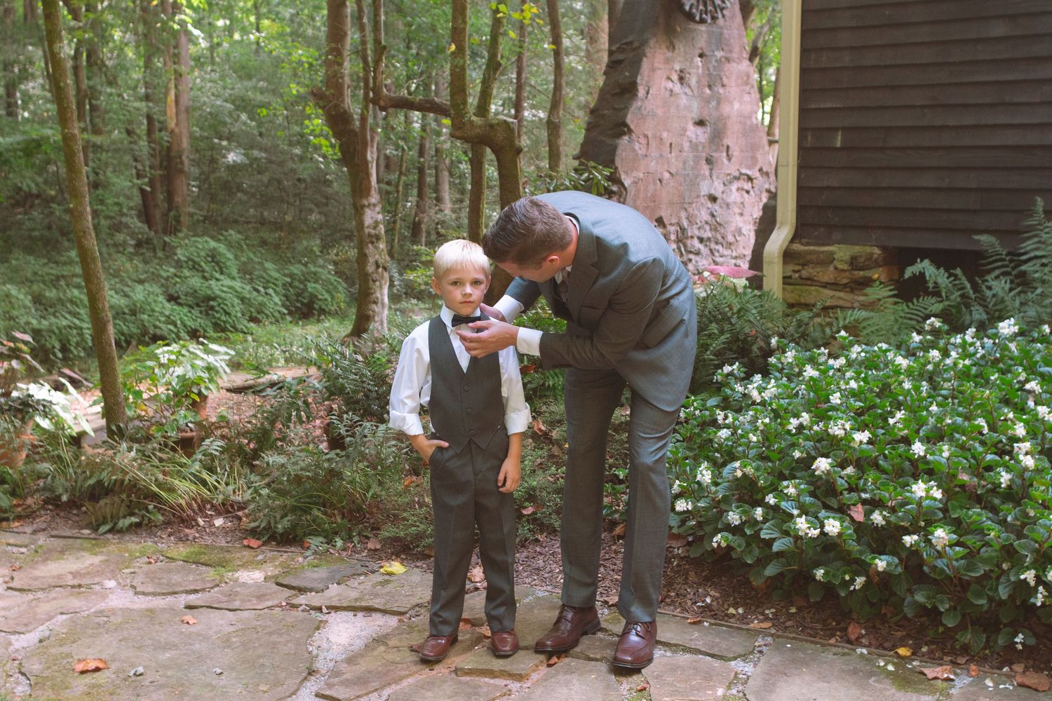 Groom adjusts young ring bearer's boutonniere on a garden stone path surrounded by greenery.