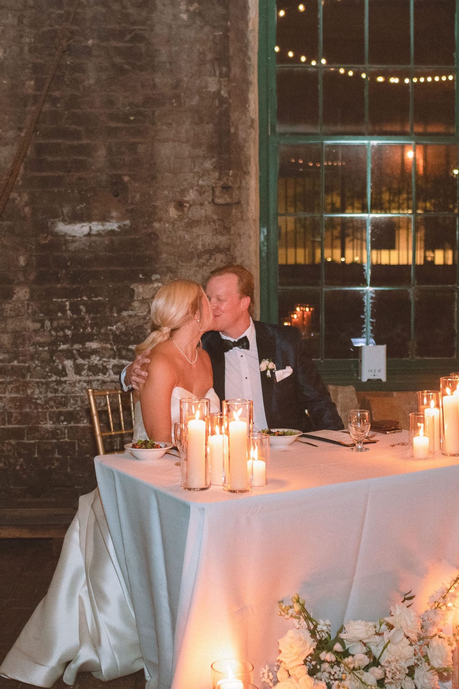 Bride and groom sharing a kiss at candlelit sweetheart table during reception.