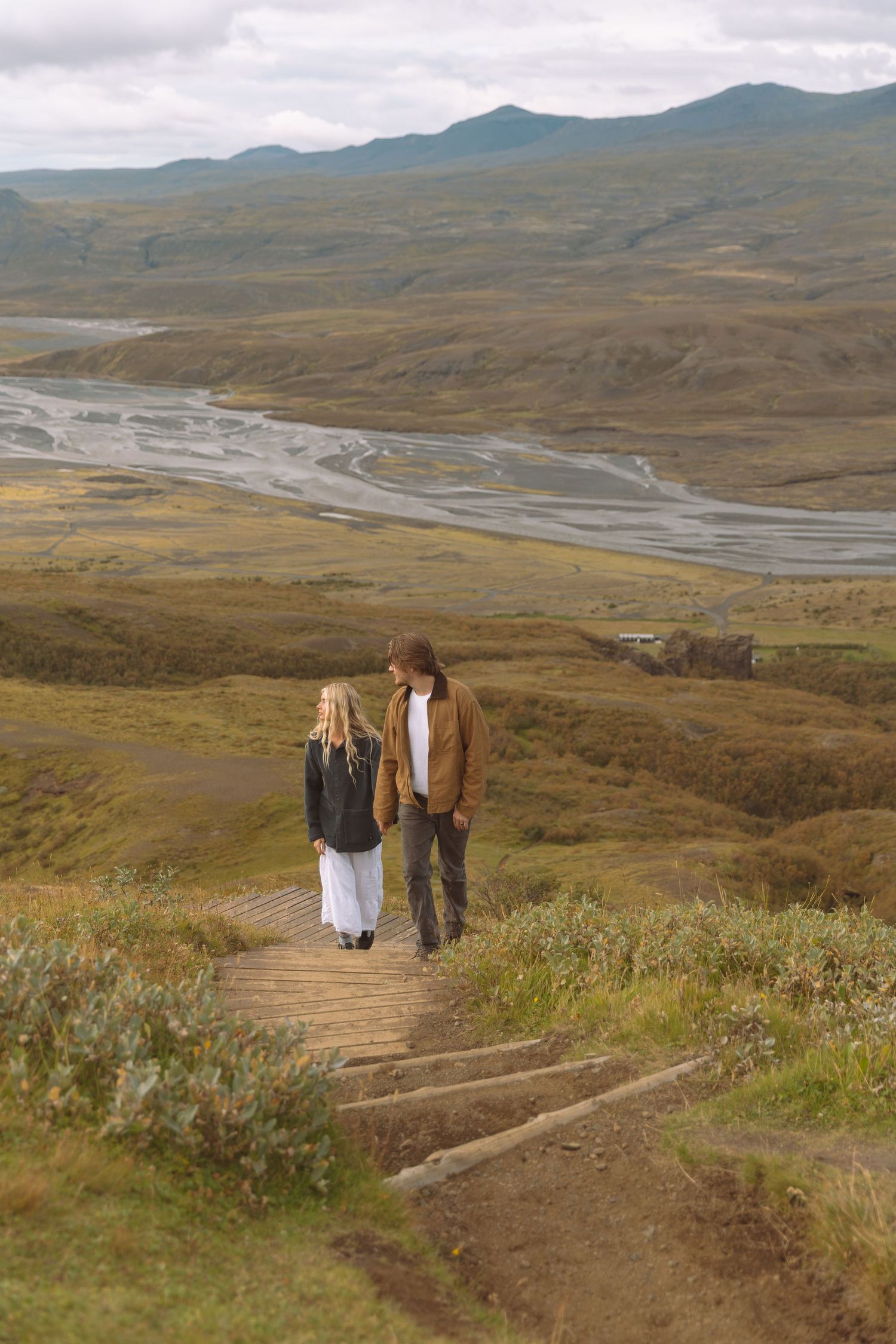 Couple walking hand in hand on a wooden path overlooking a vast green valley with a winding river and mountains.