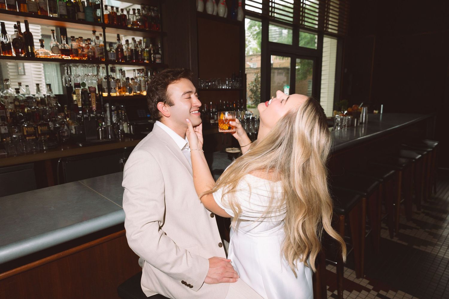 Couple laughing together at a bar, woman in white dress holding a cocktail while touching mans face.