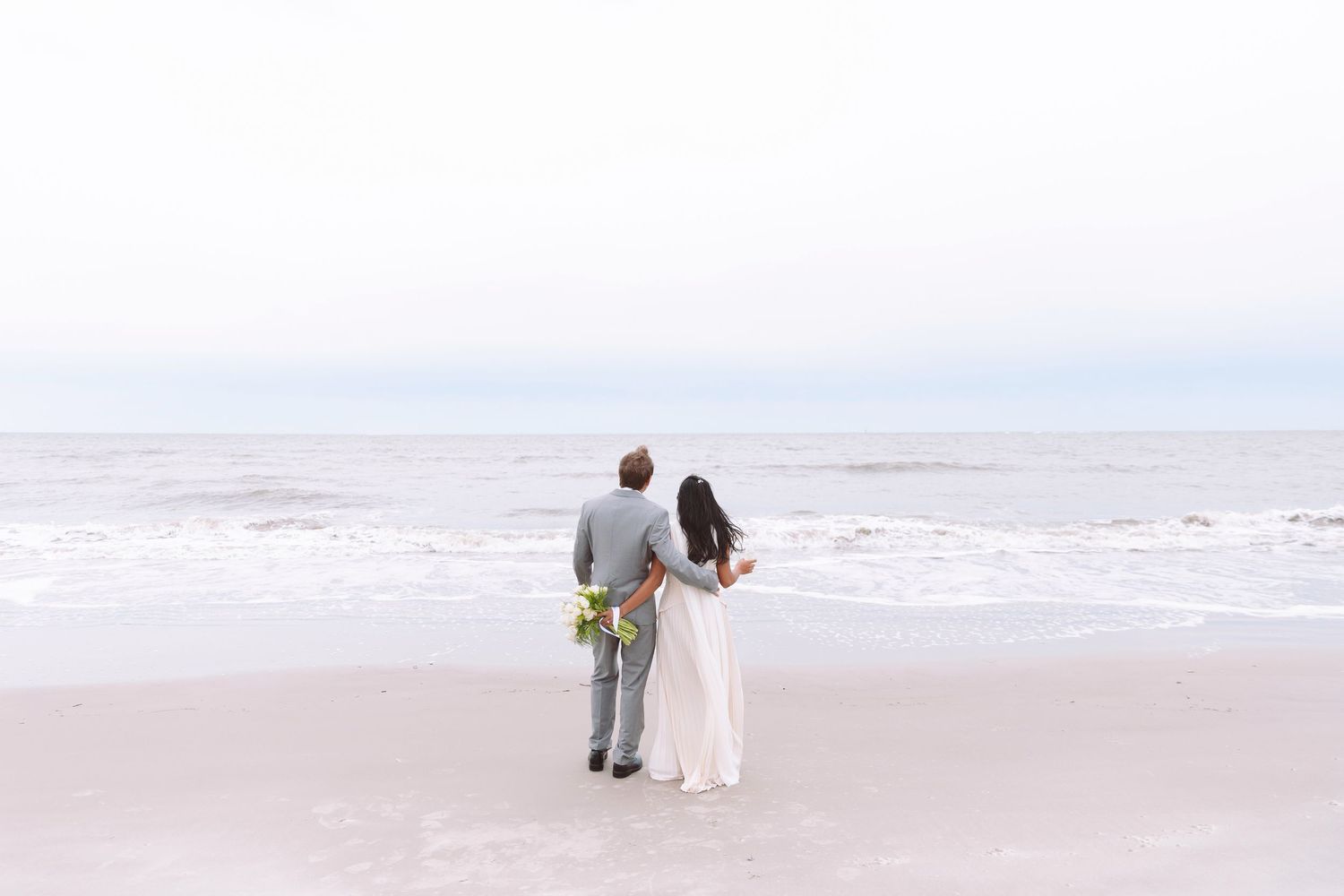 Newlywed couple stands together on empty beach gazing at ocean waves on their wedding day.