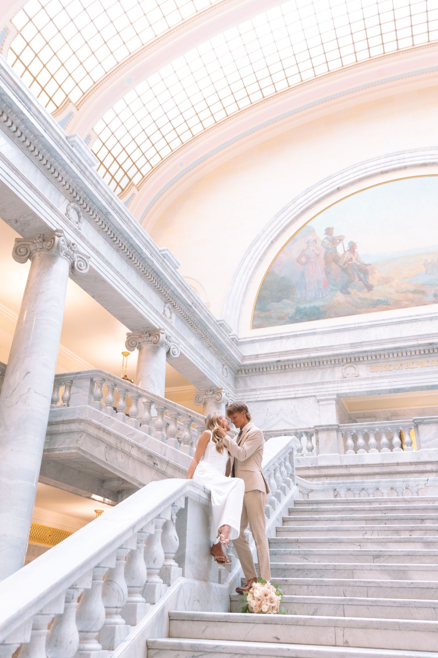 Wedding couple poses romantically on grand marble staircase beneath stunning domed ceiling with mural.