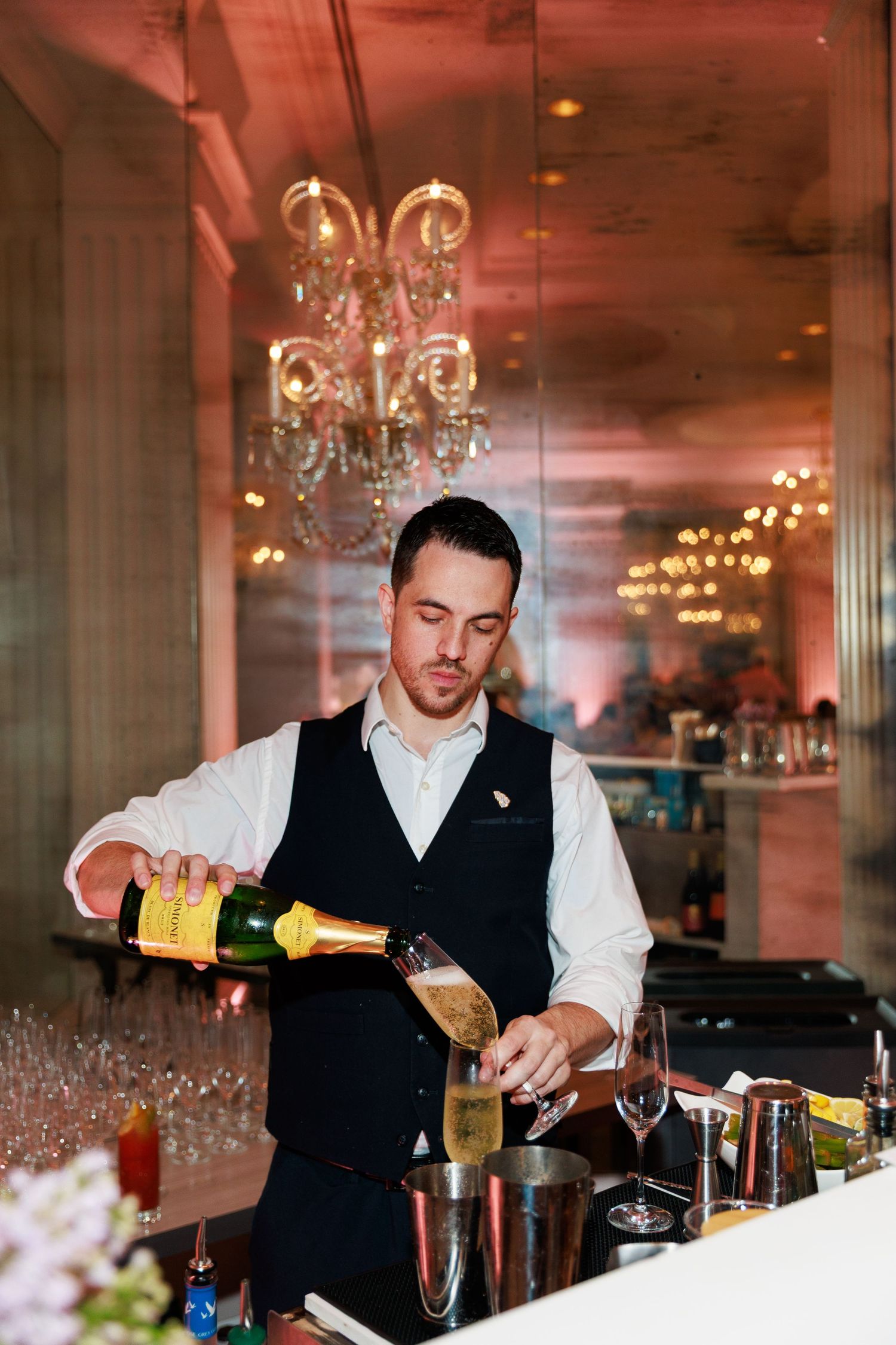 Bartender in vest pouring champagne into flute at elegant bar with crystal chandeliers.