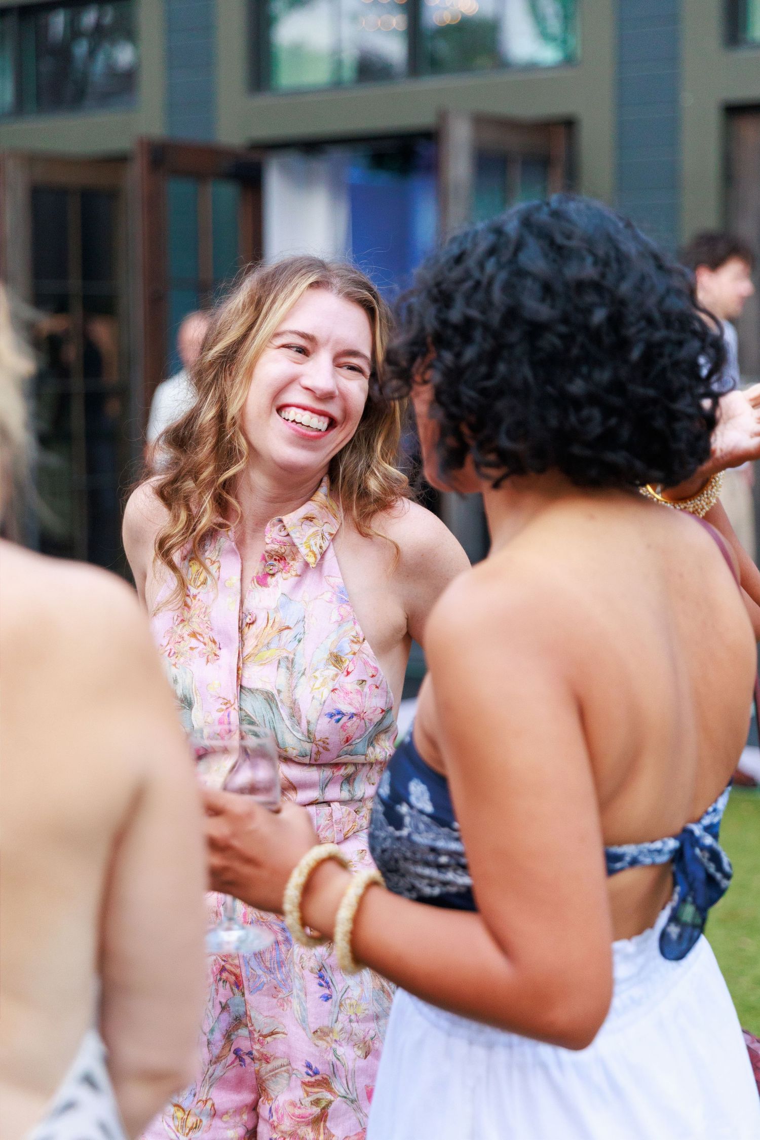 Two women laughing and socializing outdoors at a summer party, one wearing a floral pink dress.