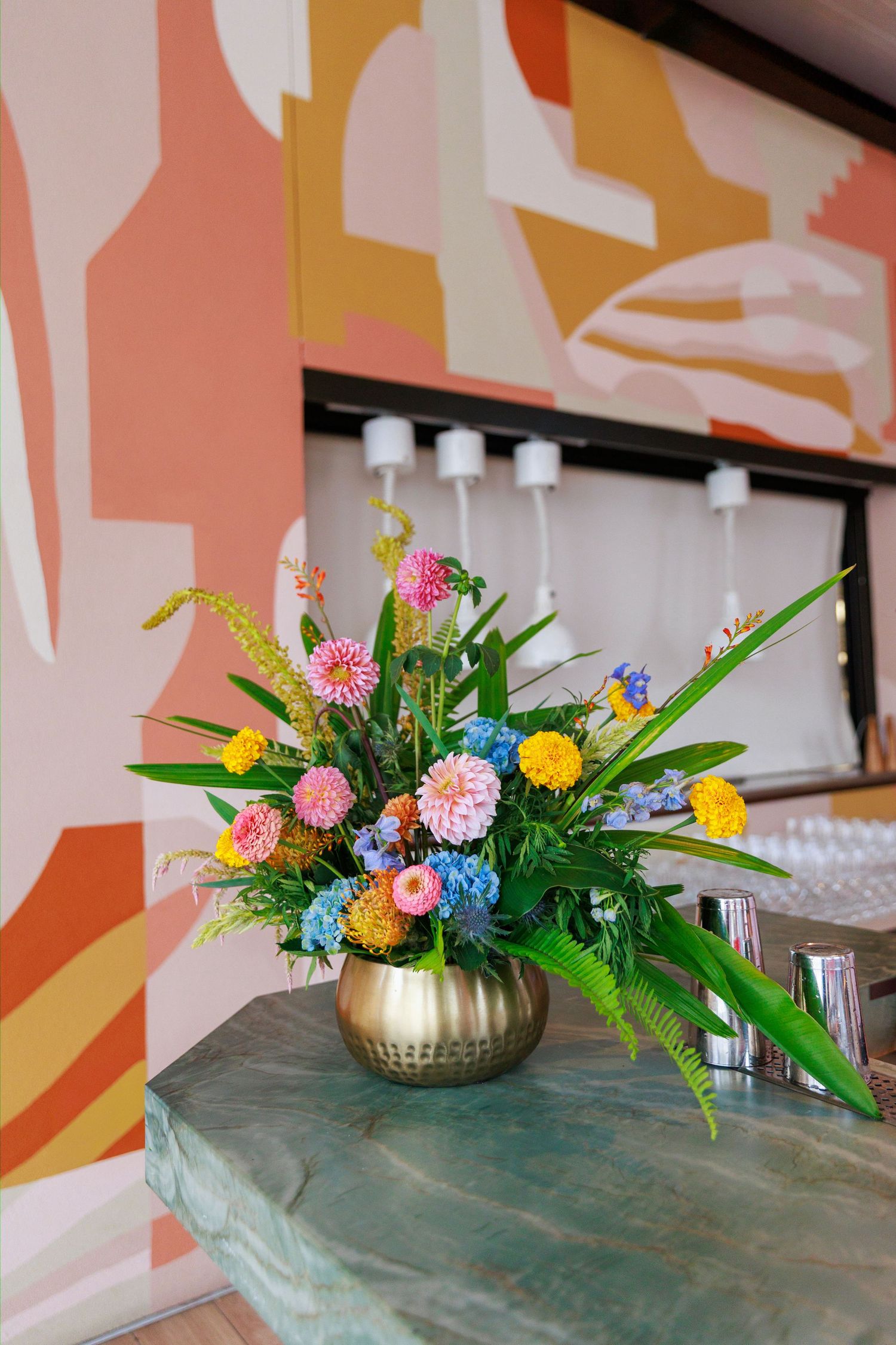 Colorful floral arrangement in gold vase with dahlias, ferns, and wildflowers on marble table against vibrant abstract mural.