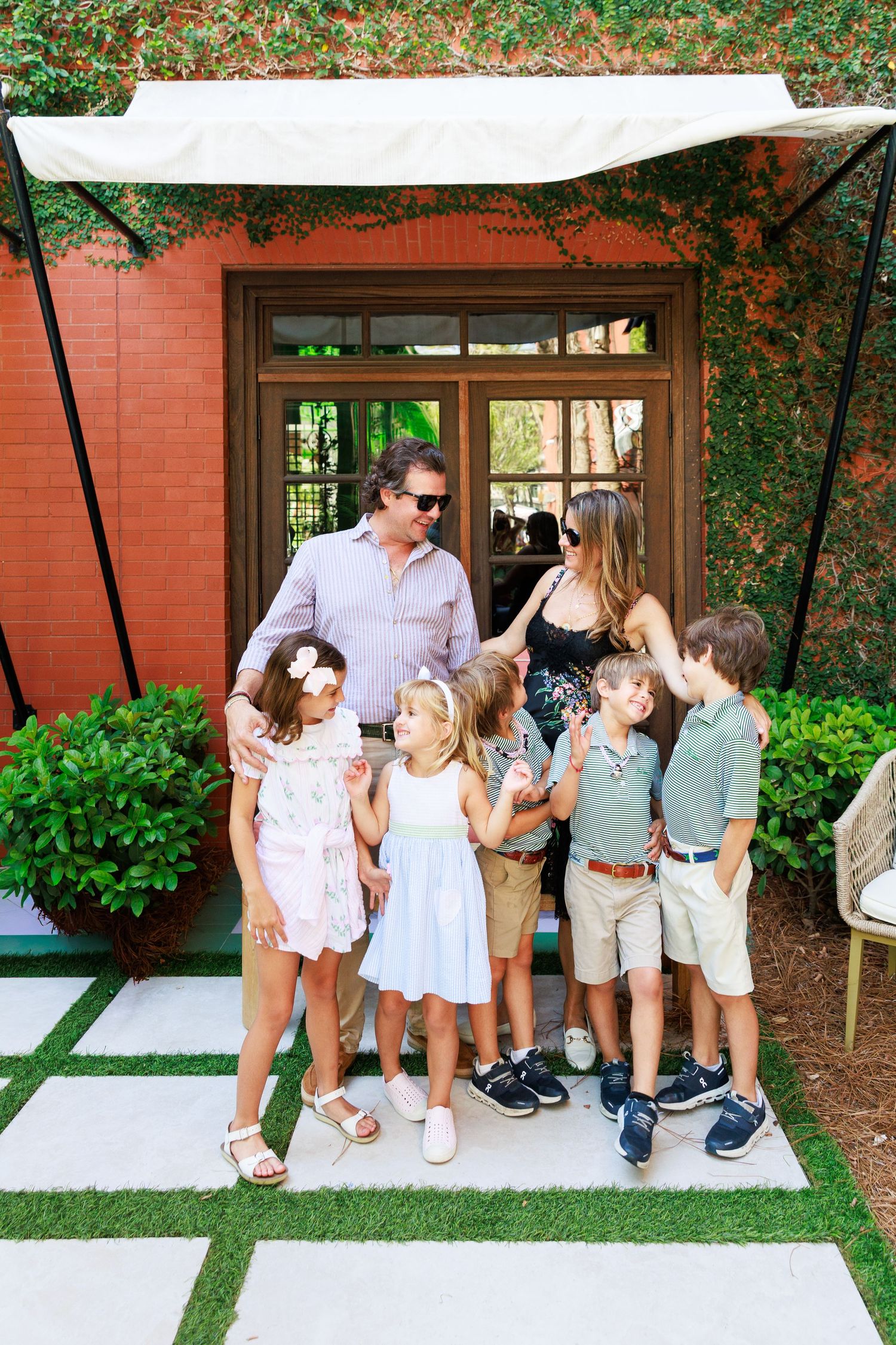 A family of seven poses outdoors in front of a red brick building with lush greenery, including two adults and five children.