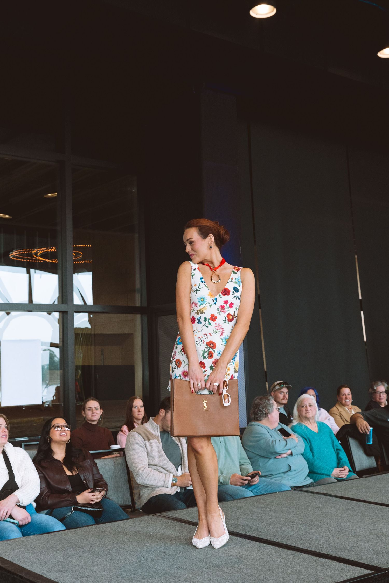 Woman modeling a floral dress with tan handbag on a runway at a fashion show with seated audience.