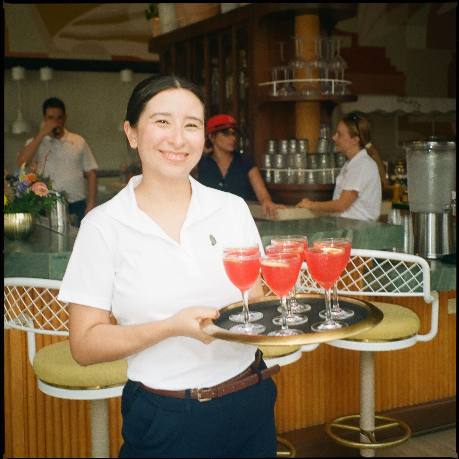 Smiling waitress in white polo shirt carrying tray of red cocktails in stemmed glasses at a bar.