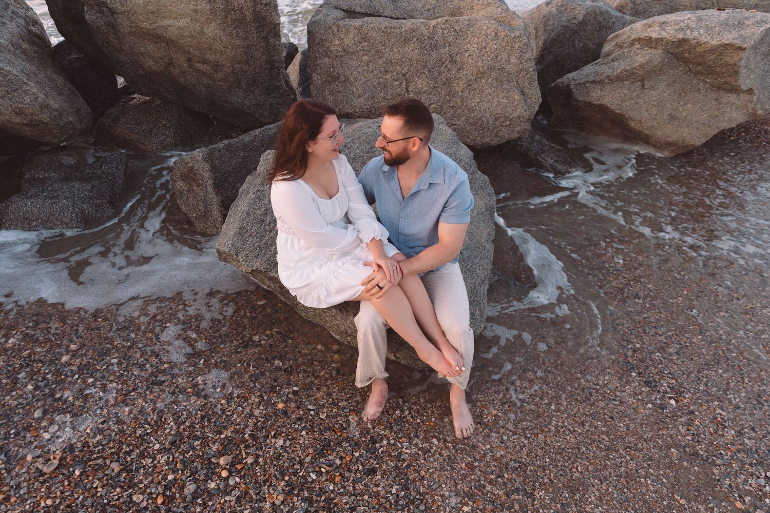 Couple sitting together on rocky beach shore, smiling at each other during golden hour sunset.
