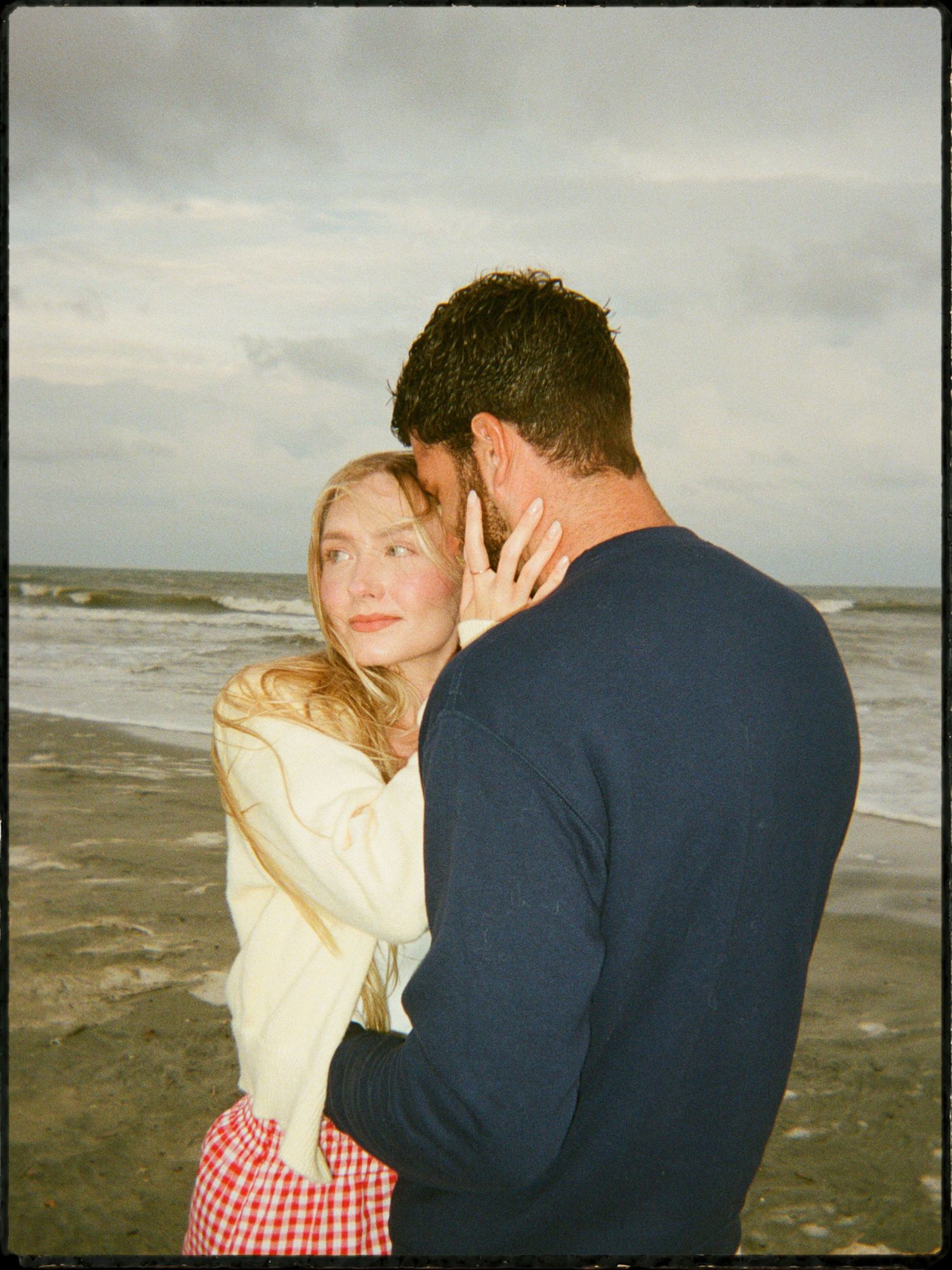 Couple sharing a tender moment on a stormy beach, woman in cream sweater and plaid skirt touching mans face.