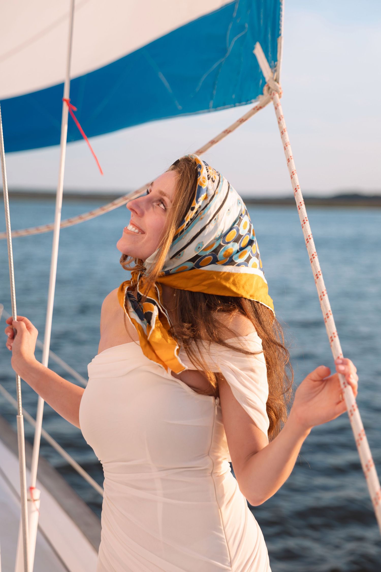 Happy woman in white dress and colorful headscarf holding sailboat ropes on a sunny day on the water.