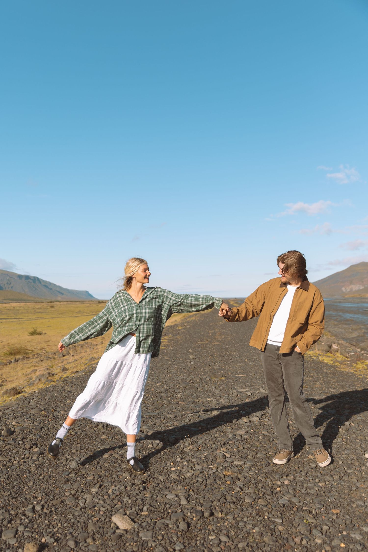 Happy couple holding hands on a scenic gravel road with mountains and blue sky.
