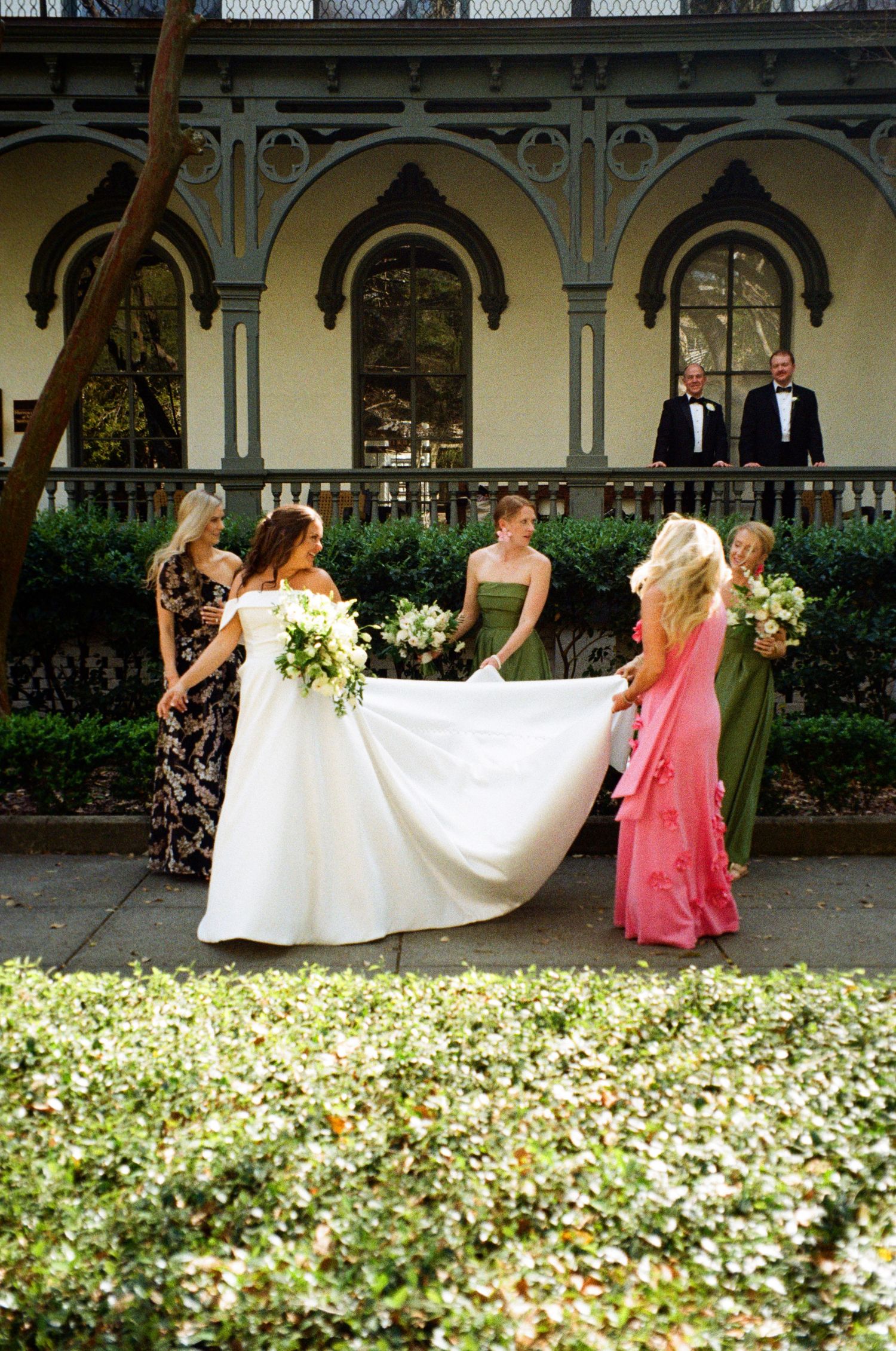 Bridesmaids in colorful gowns help bride with white dress train outside elegant historic venue.