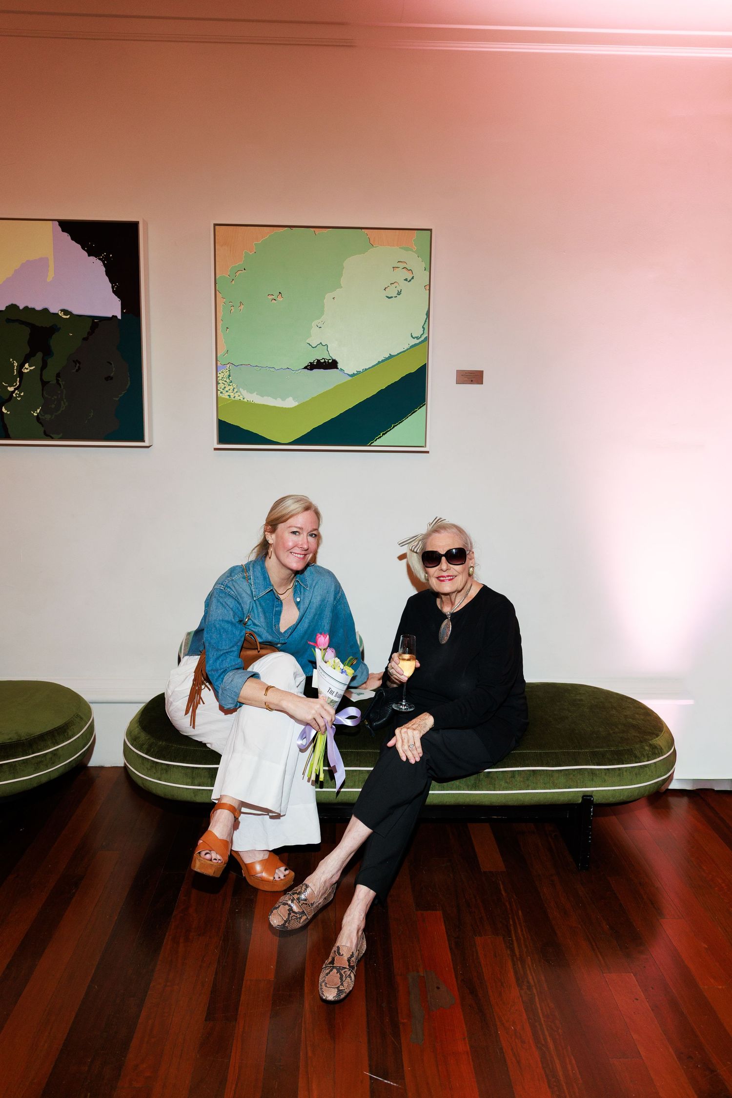 Two women sitting on a green velvet sofa at an art gallery, smiling, with colorful paintings on the wall behind them.