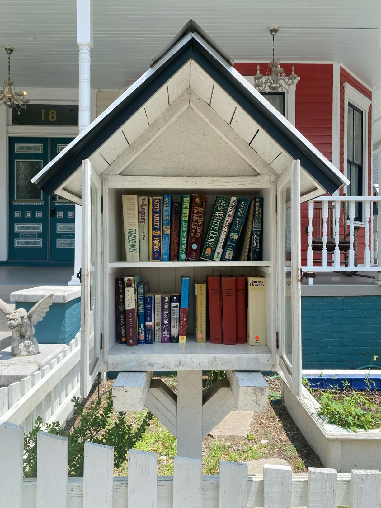 White Little Free Library filled with colorful books in front of a porch.