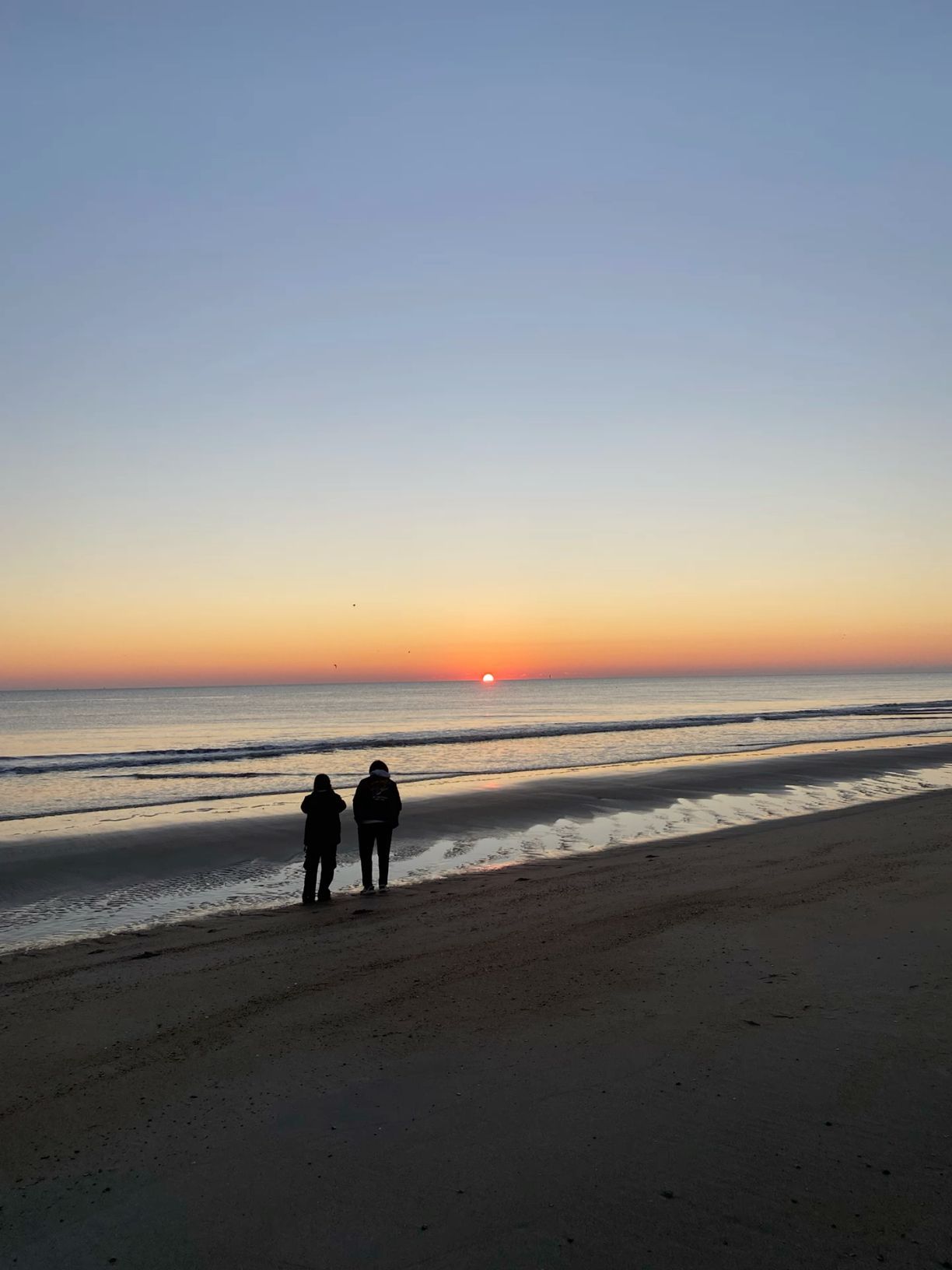 Two silhouetted figures stand on a sandy beach watching a vibrant orange and red sunset over calm ocean waves.