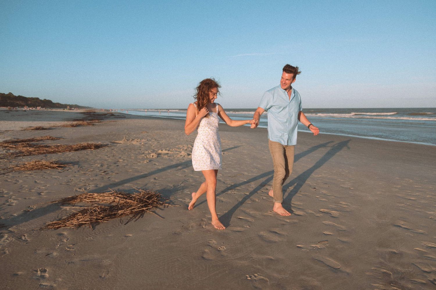 Happy couple holding hands and walking along a sandy beach at golden hour near the ocean.