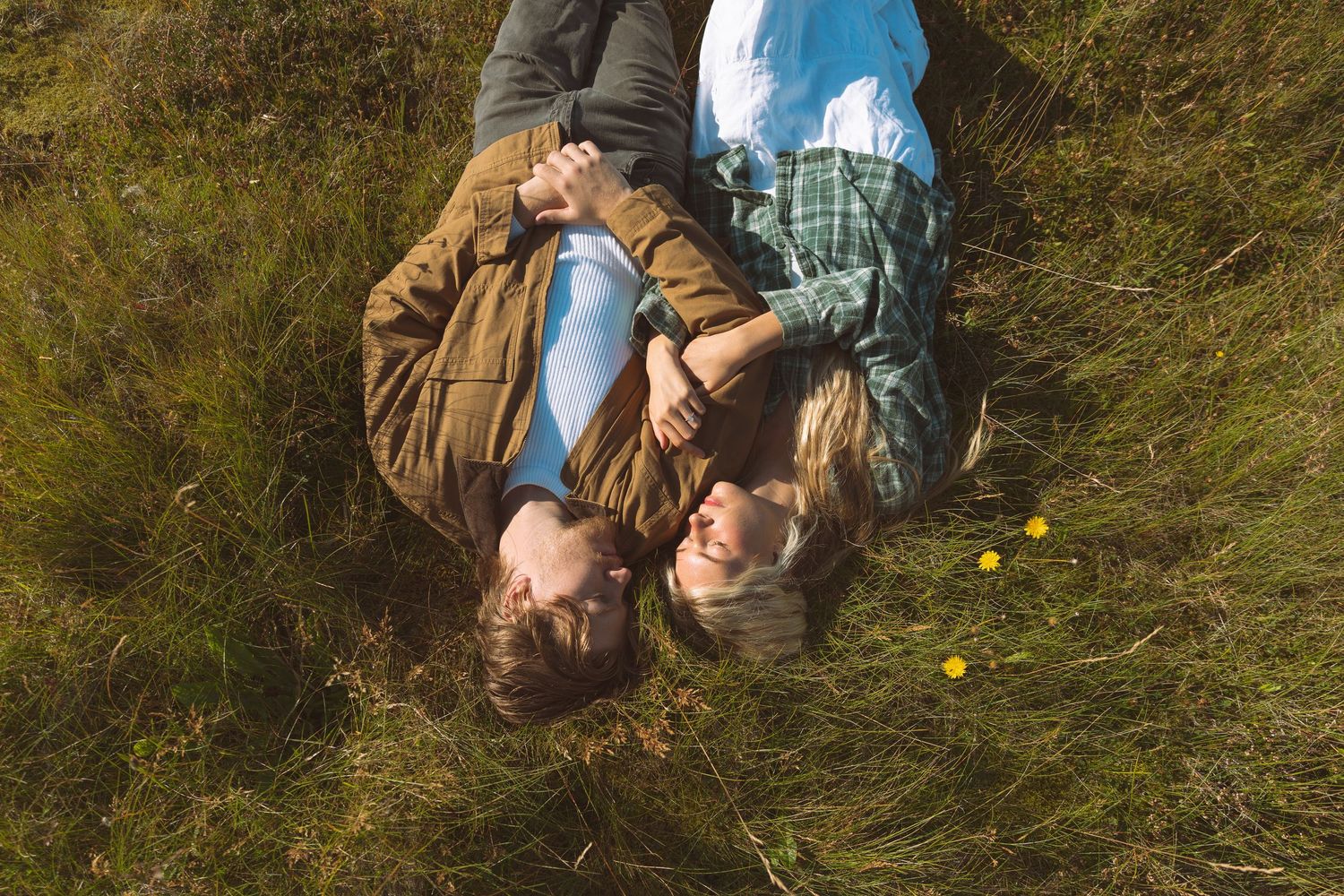 Couple lies together in green grass holding hands during a romantic outdoor engagement session.