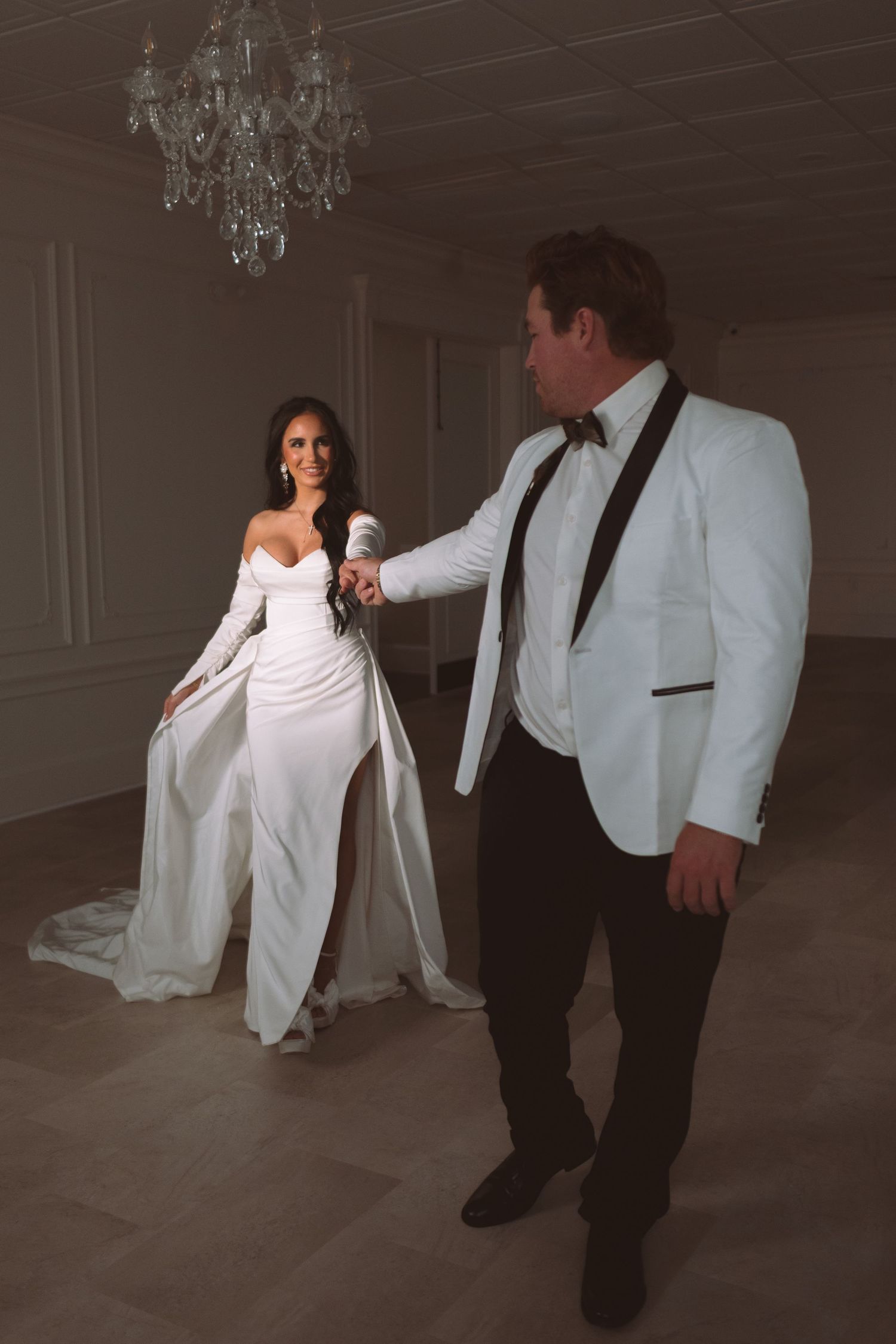Elegant bride in a white off-shoulder gown holds hands with groom in white tuxedo under a crystal chandelier.