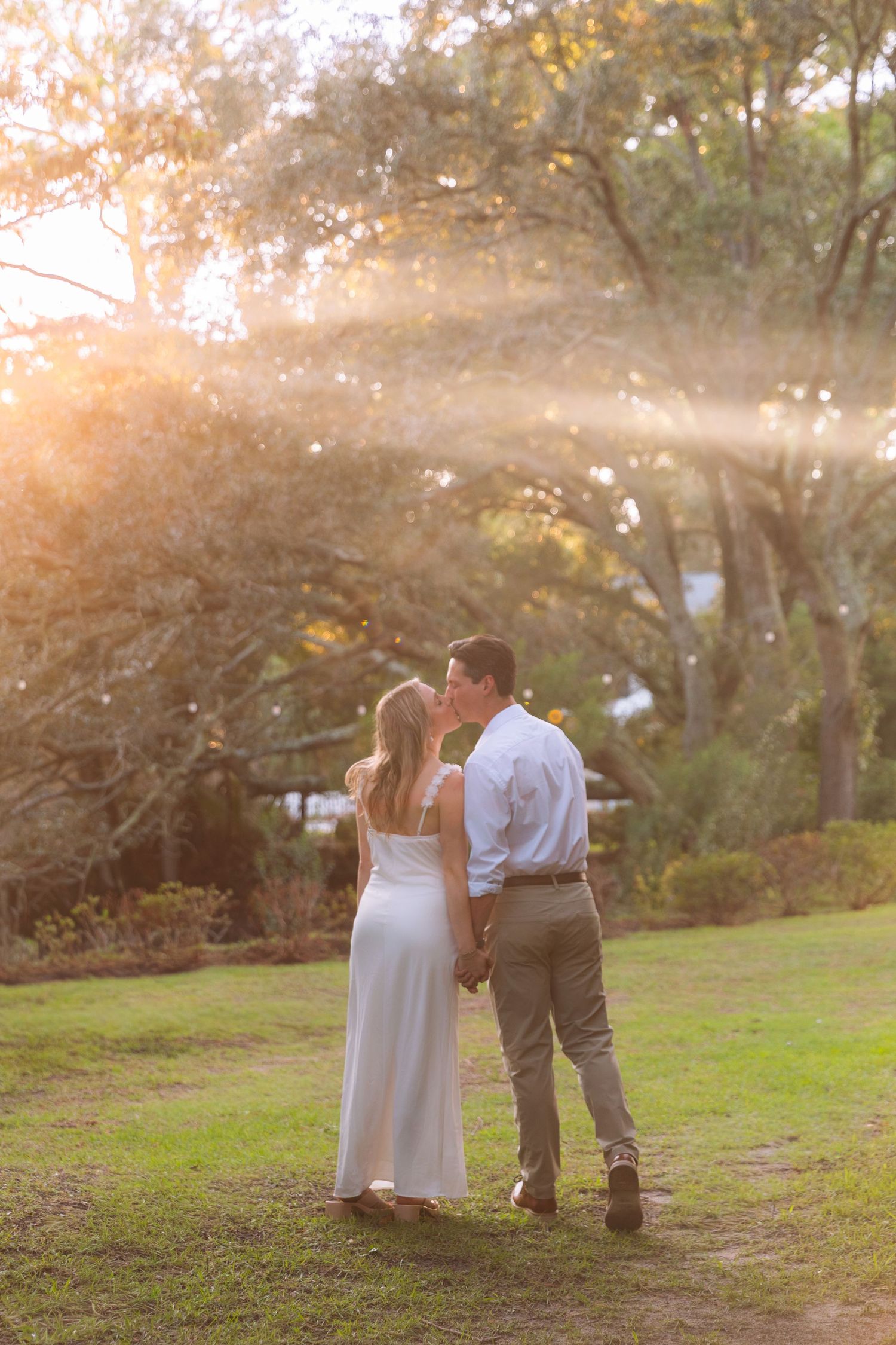 Couple walking hand-in-hand through golden sunset light in lush green park, woman in white dress.