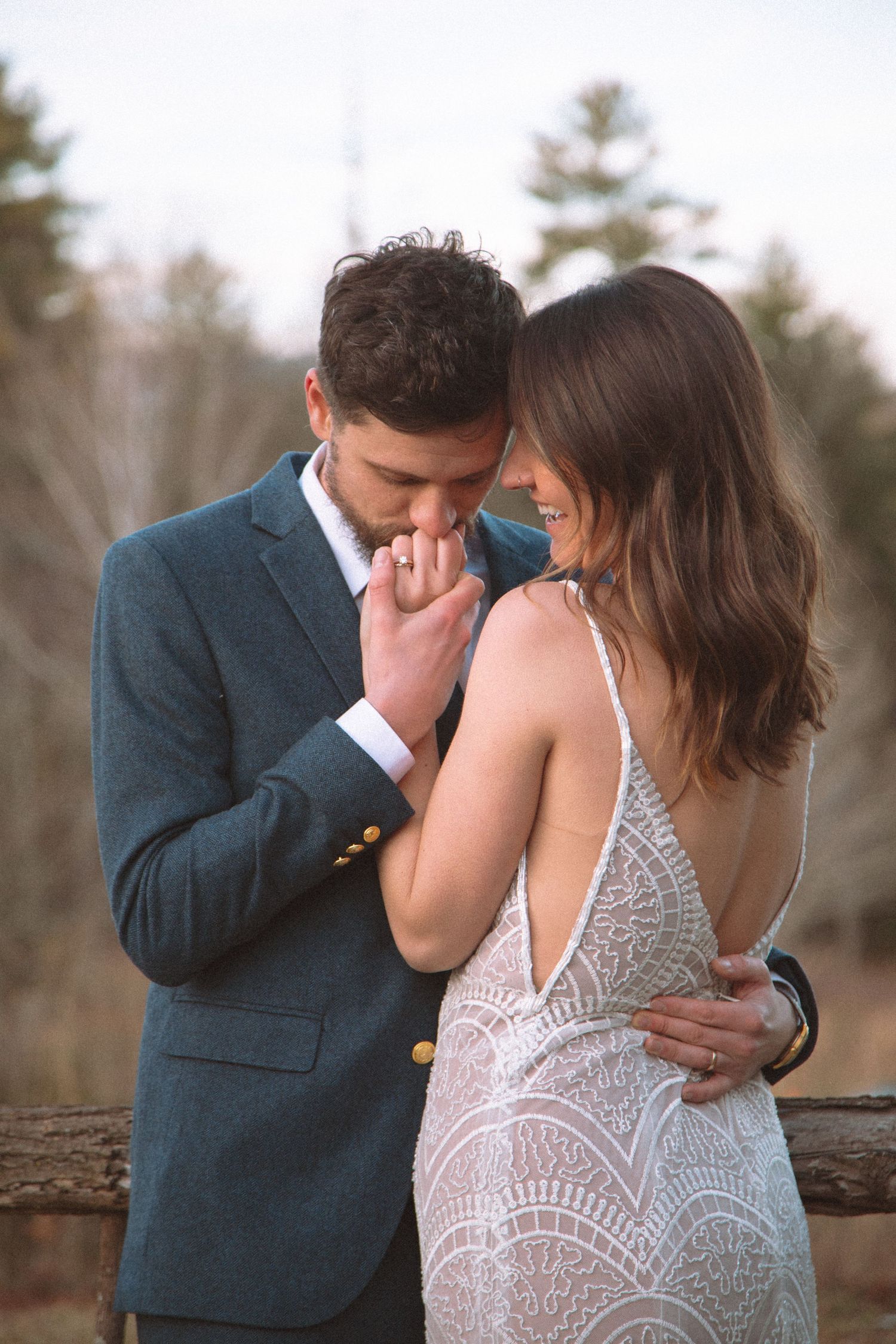 A groom in a blue suit kisses his bride's hand as she smiles in a lace backless wedding dress outdoors.
