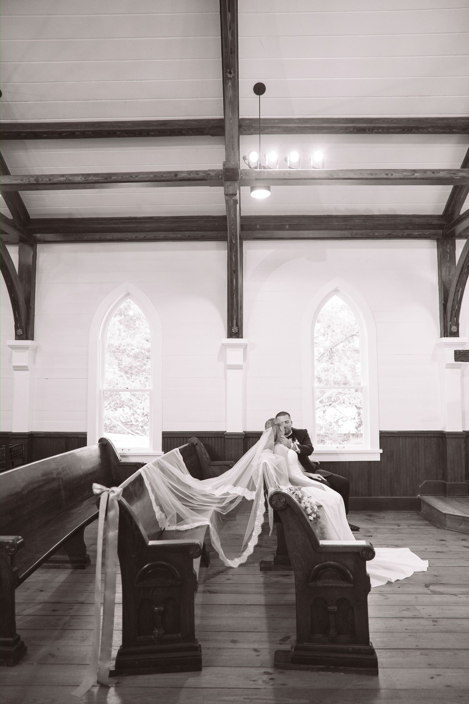 Black and white photo of bride and groom sitting in church pews with flowing veil draped across aisle.