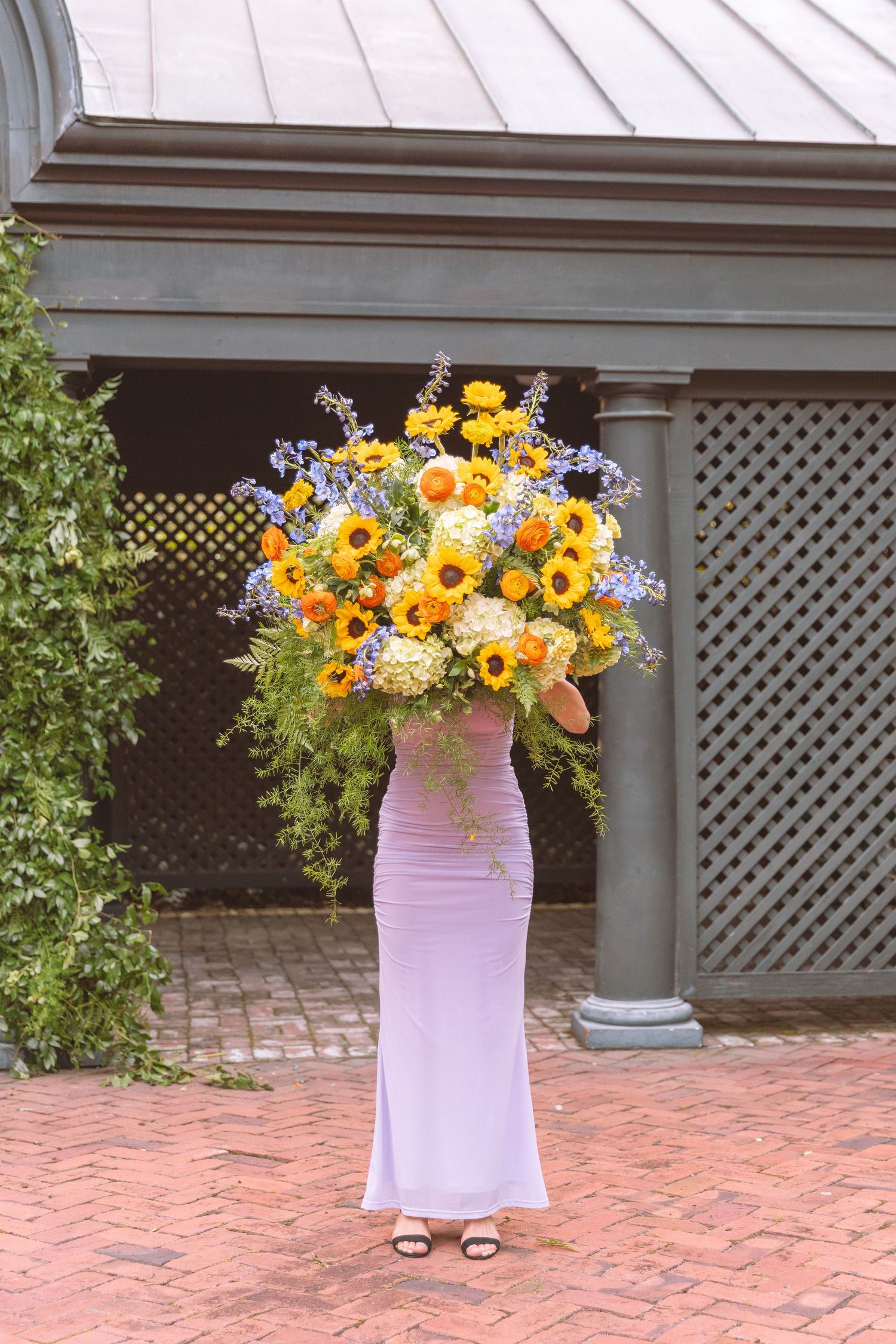 Person in lavender dress carries an oversized sunflower and wildflower bouquet obscuring their face on a brick patio.