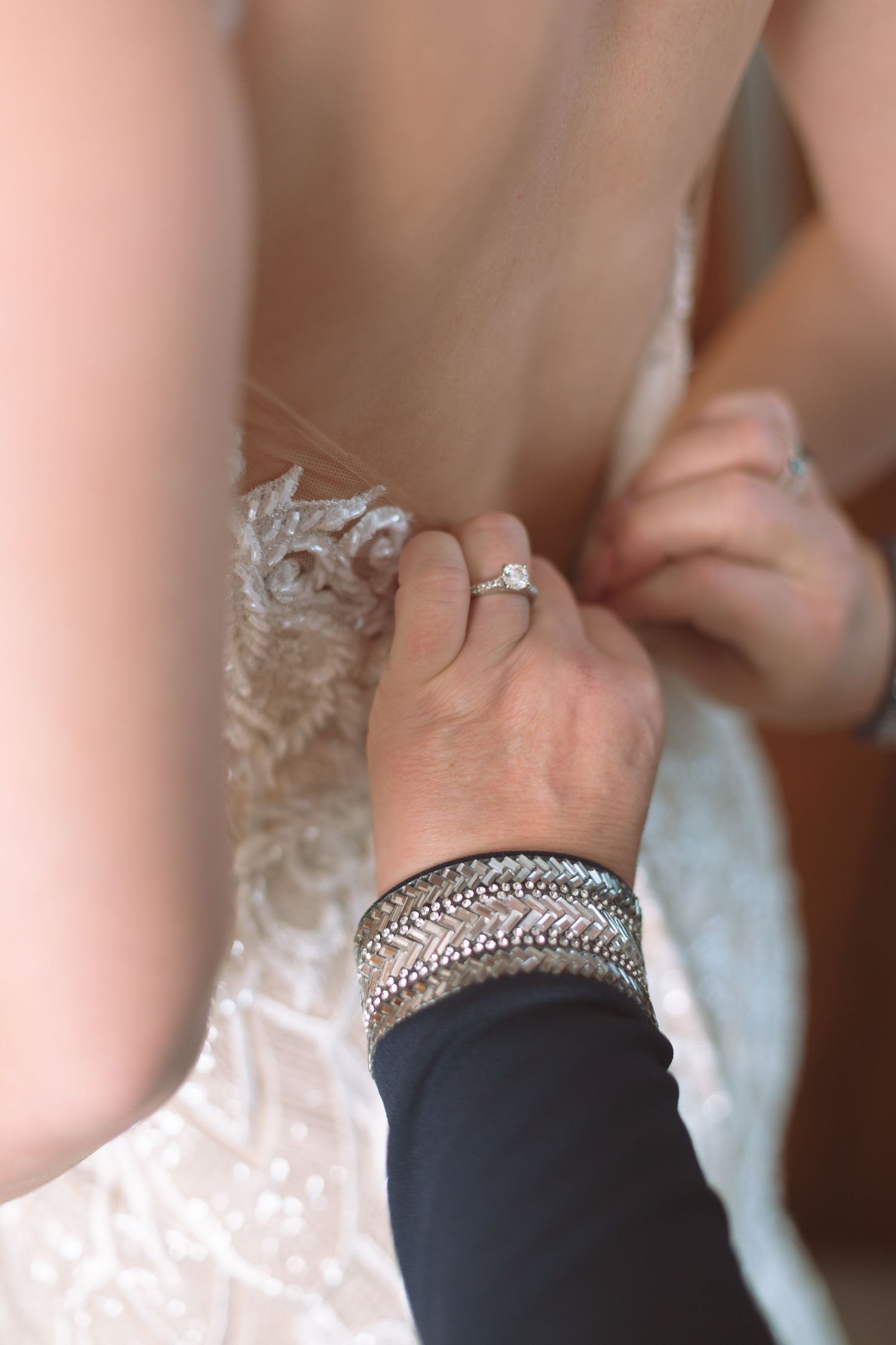Close-up of hands fastening buttons on back of intricate lace wedding dress with diamond ring.