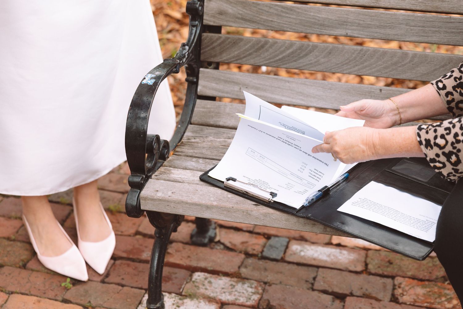 Bride in white heels signing wedding documents on a wooden bench with a clipboard outdoors.
