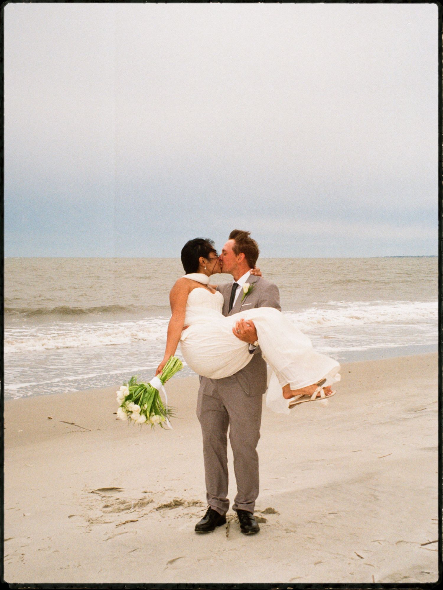 Groom lifts bride in arms for romantic beach kiss while holding white calla lily bouquet.