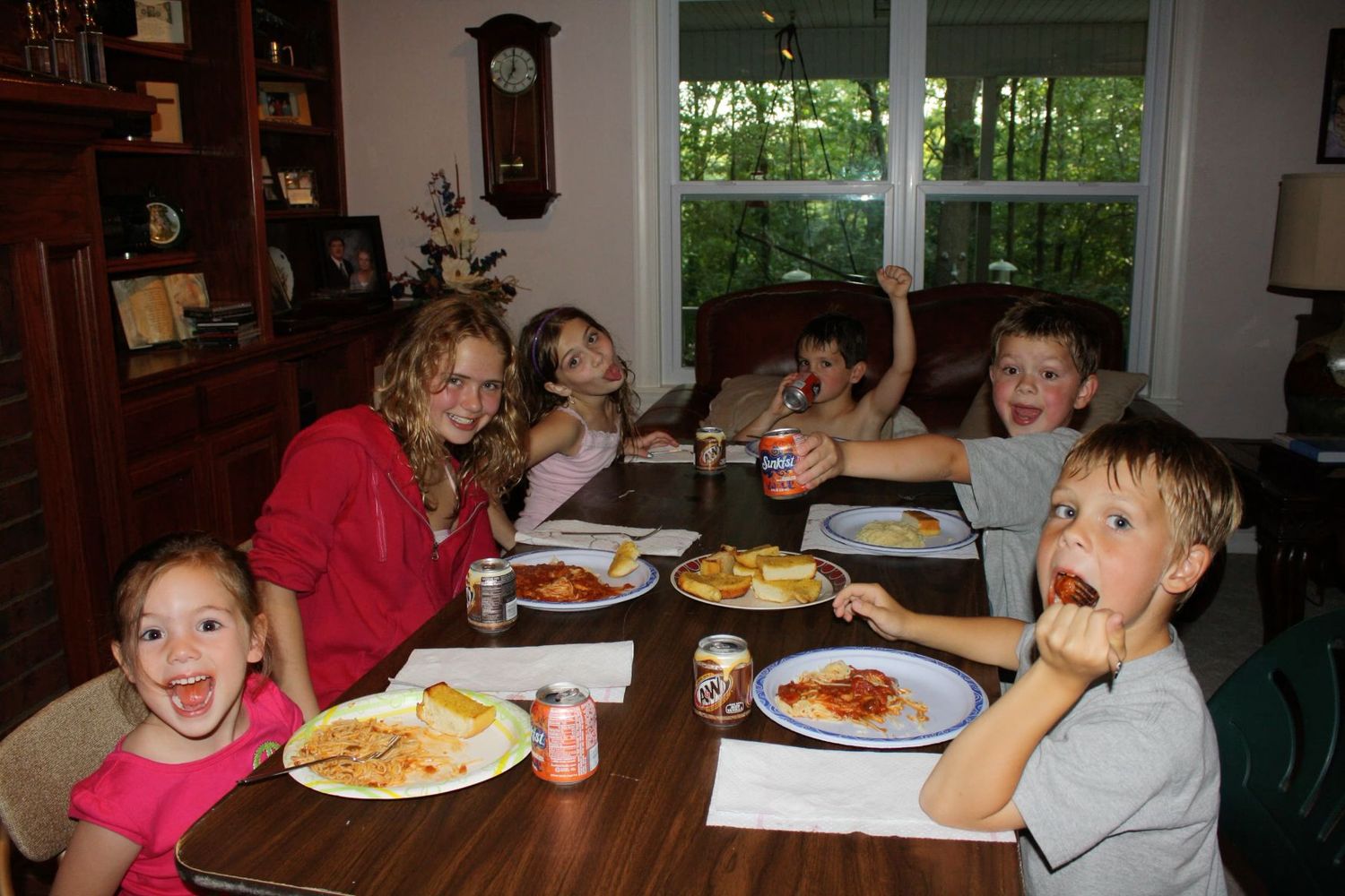 Group of children and an adult smiling while eating pasta dinner together at a dining room table.