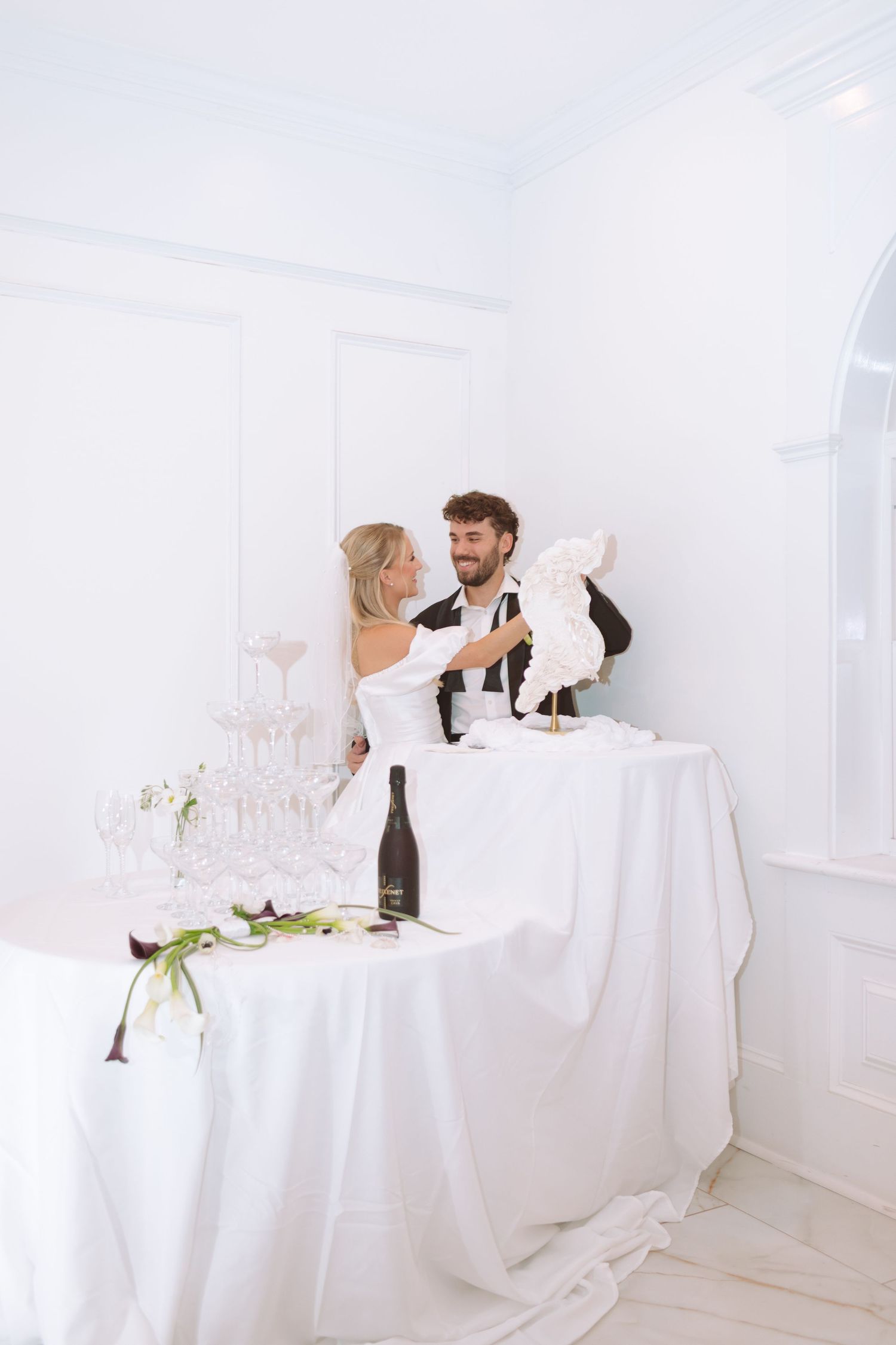 Bride and groom admiring a floral wedding sculpture beside a champagne tower in an elegant white venue.