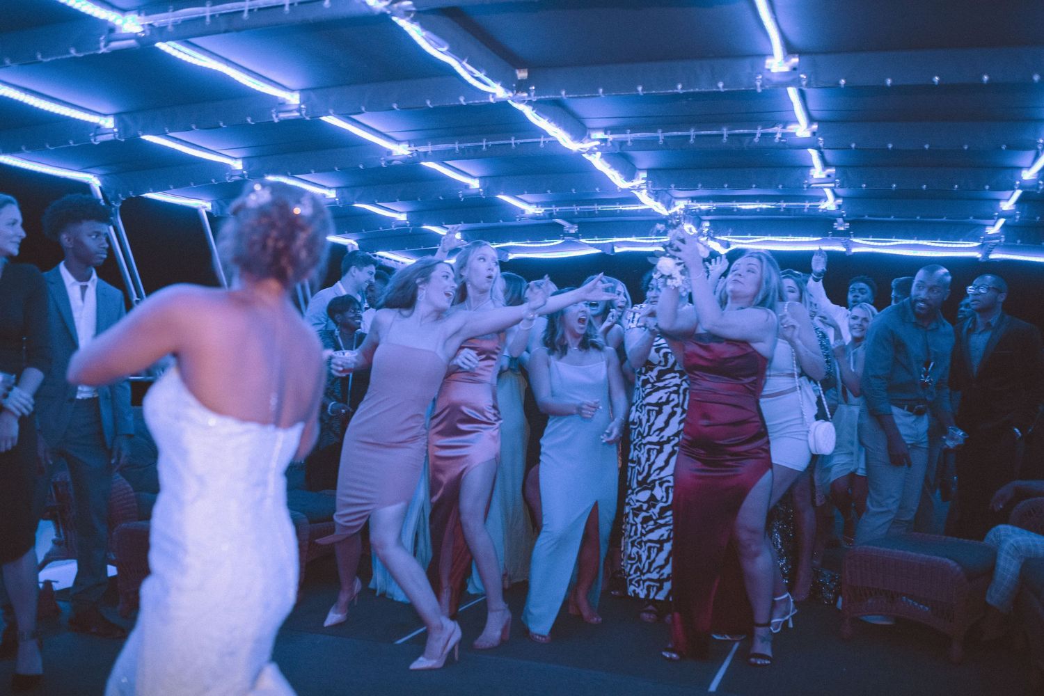 Bride facing bridesmaids in colorful gowns during bouquet toss at wedding reception.
