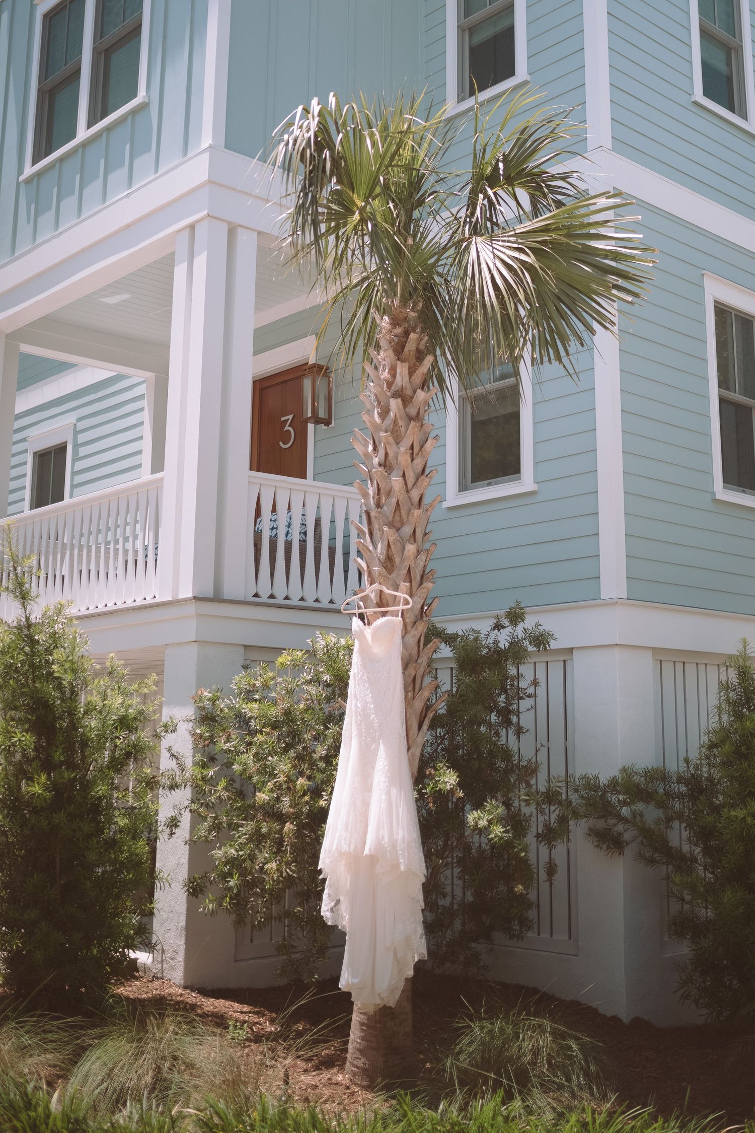 White lace wedding dress hanging from a palm tree outside a light blue coastal beach house.