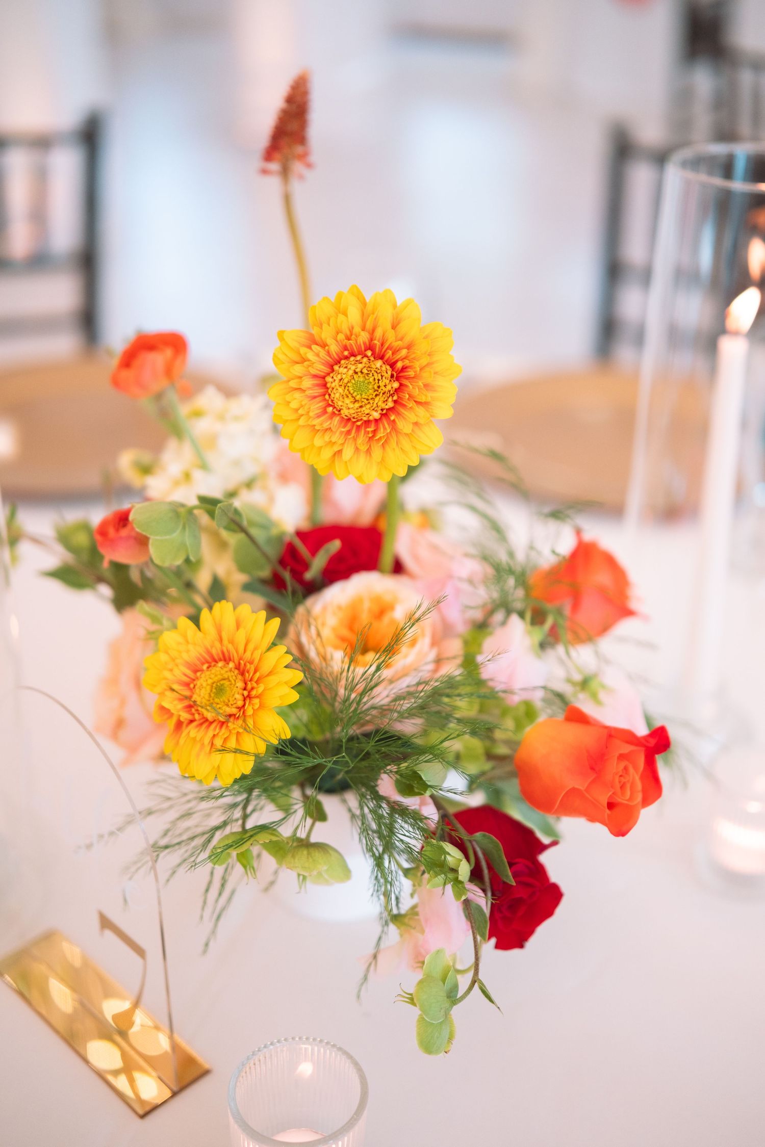 Vibrant wedding table centerpiece featuring yellow gerbera daisies, orange and red roses in white vase.