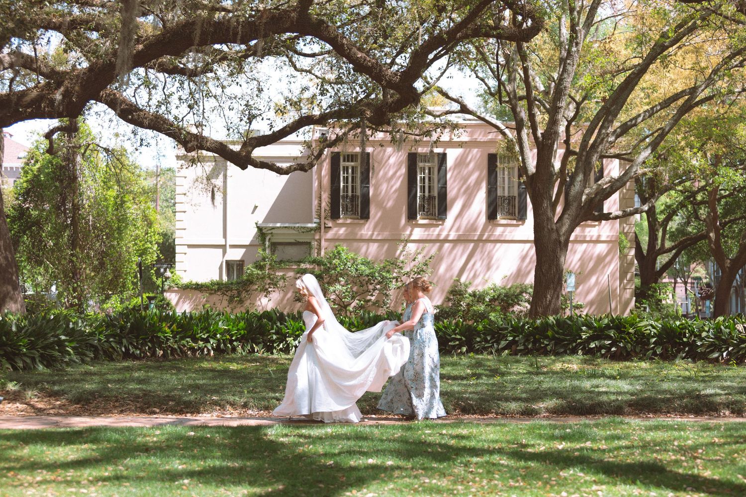 Bride and groom twirling in sunlit garden beneath Spanish moss-draped trees near pink building.