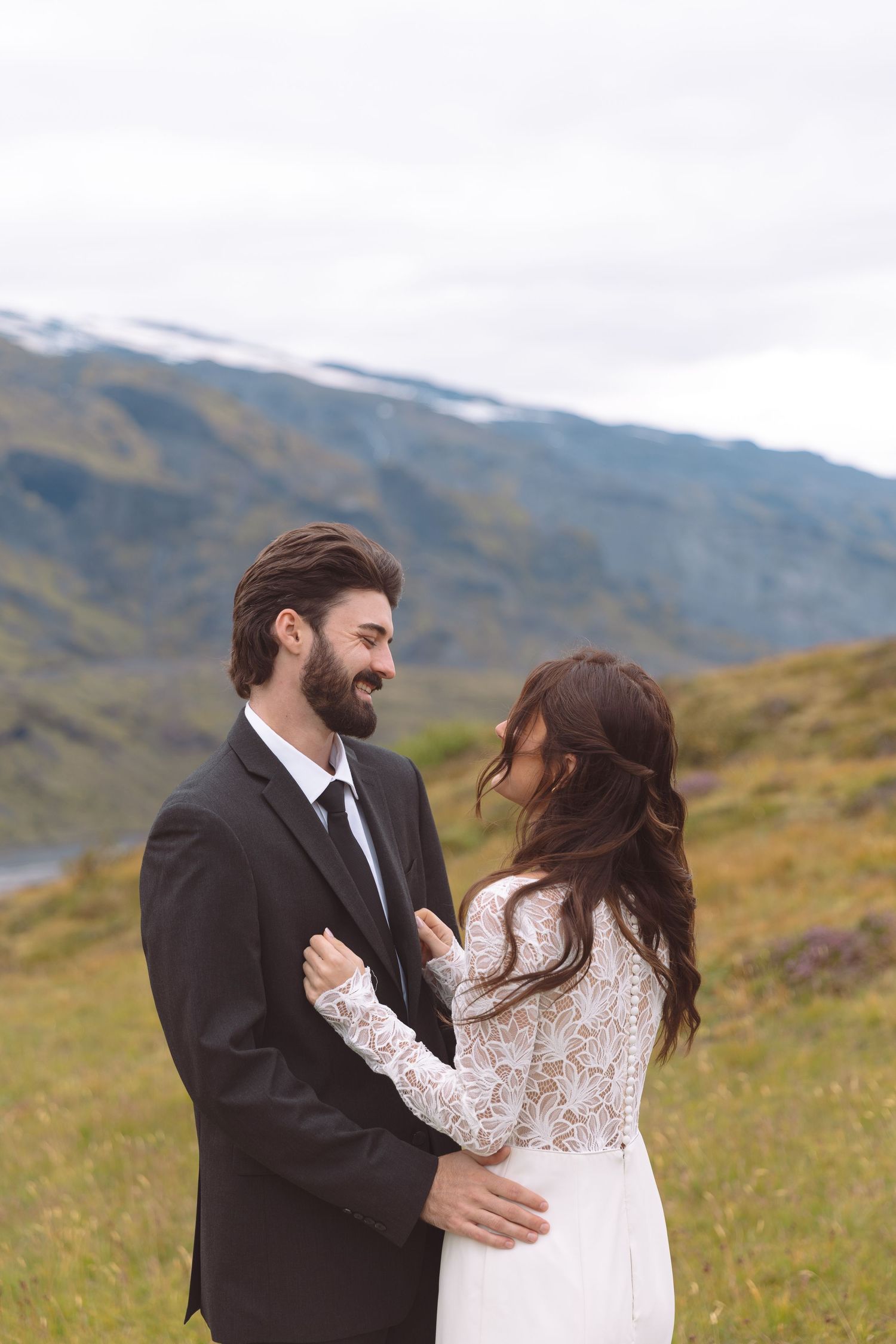 Laughing groom in black suit holds bride in lace wedding dress in a scenic mountain meadow setting.