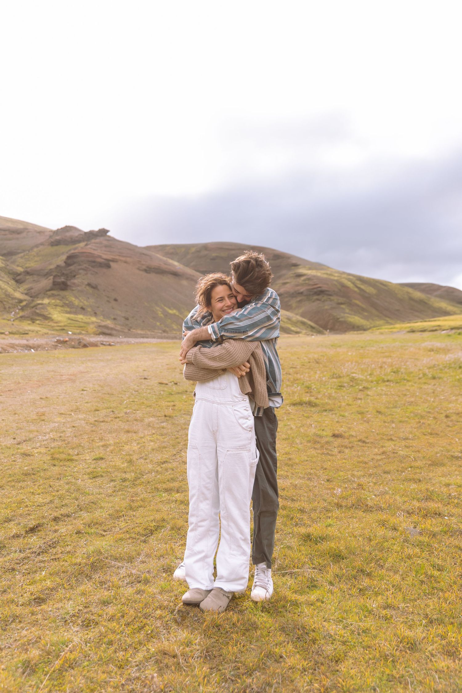 Romantic couple sharing a tender embrace in a vast green highland meadow under overcast skies.
