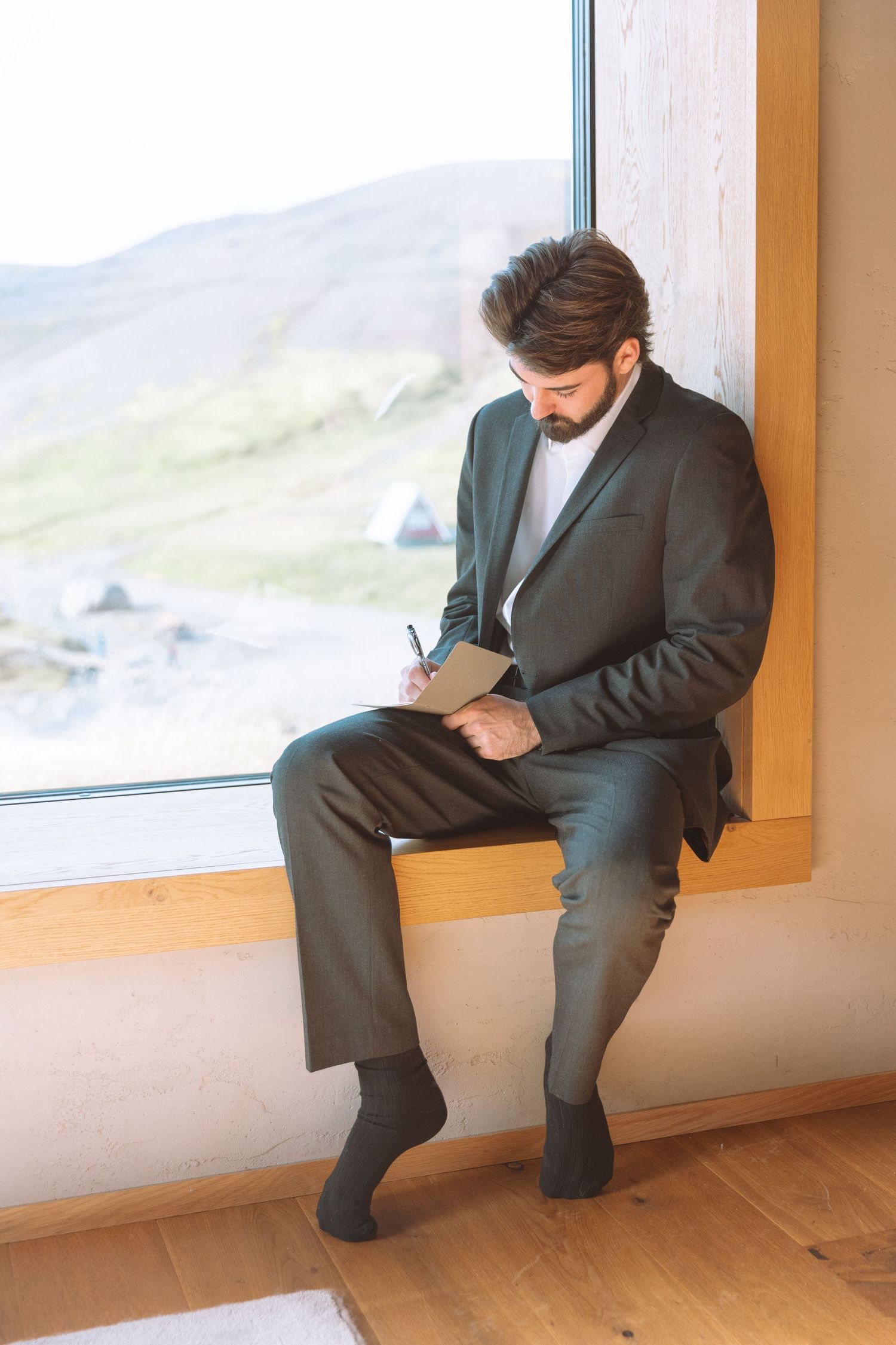 A bearded groom in a dark suit writes in a small notebook while seated by a large window.