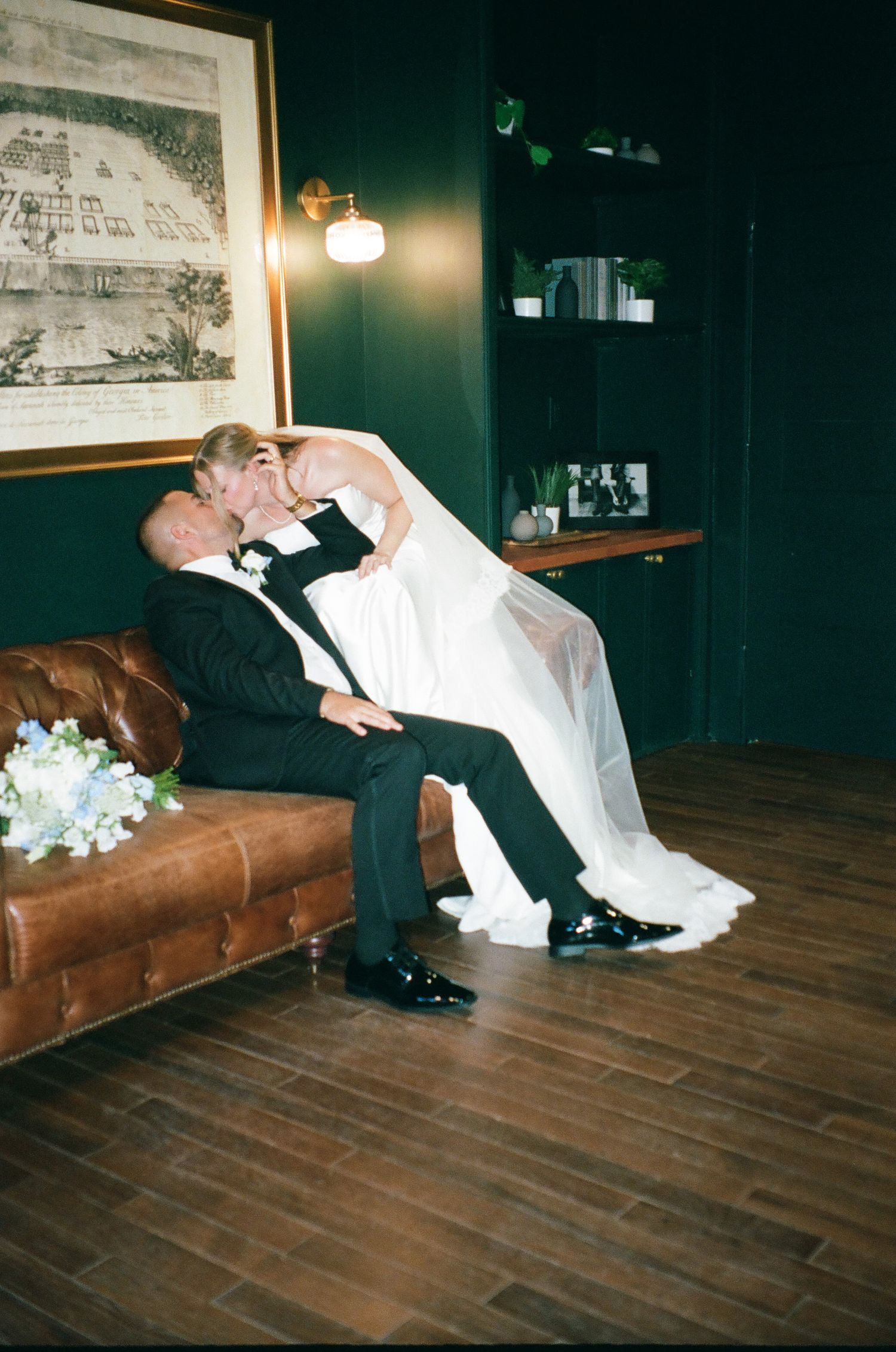 A bride in a white gown and groom in a tuxedo share a romantic kiss on a leather sofa in an elegant dark-walled room.