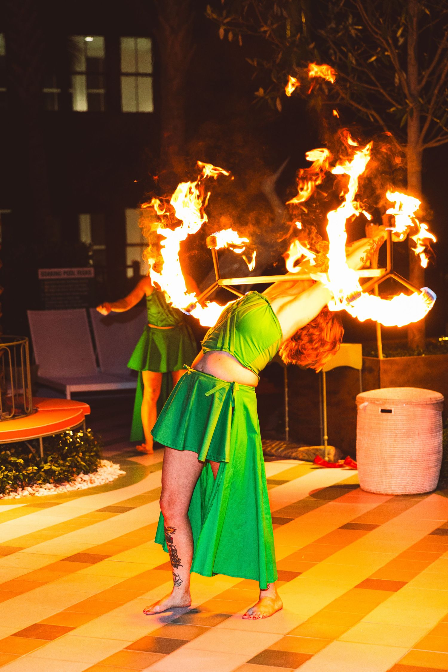 Fire dancer in green costume performs backbend with flaming hoop at night outdoor event.