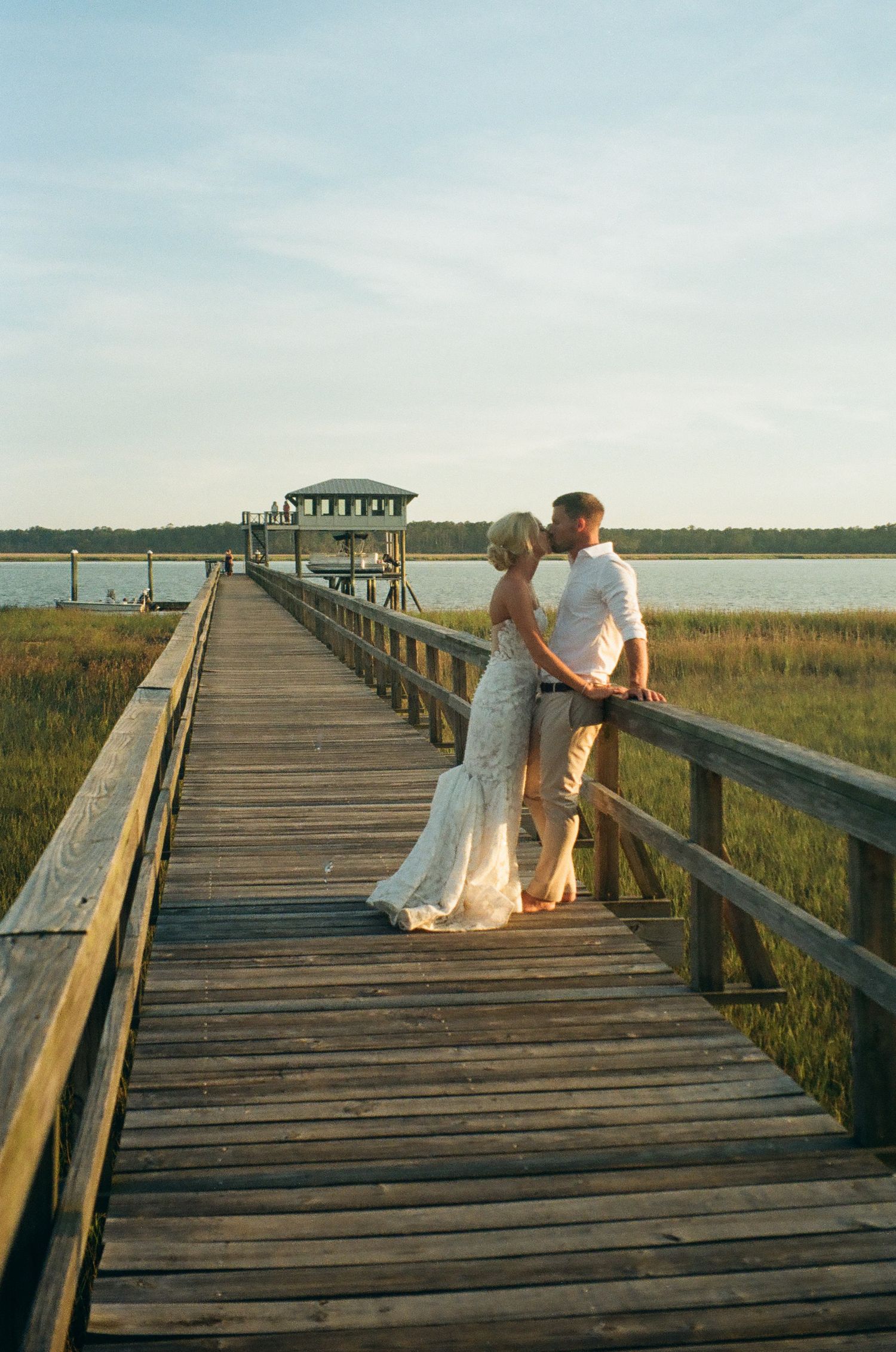 Bride and groom share a romantic kiss on a wooden boardwalk overlooking a serene coastal marsh at sunset.